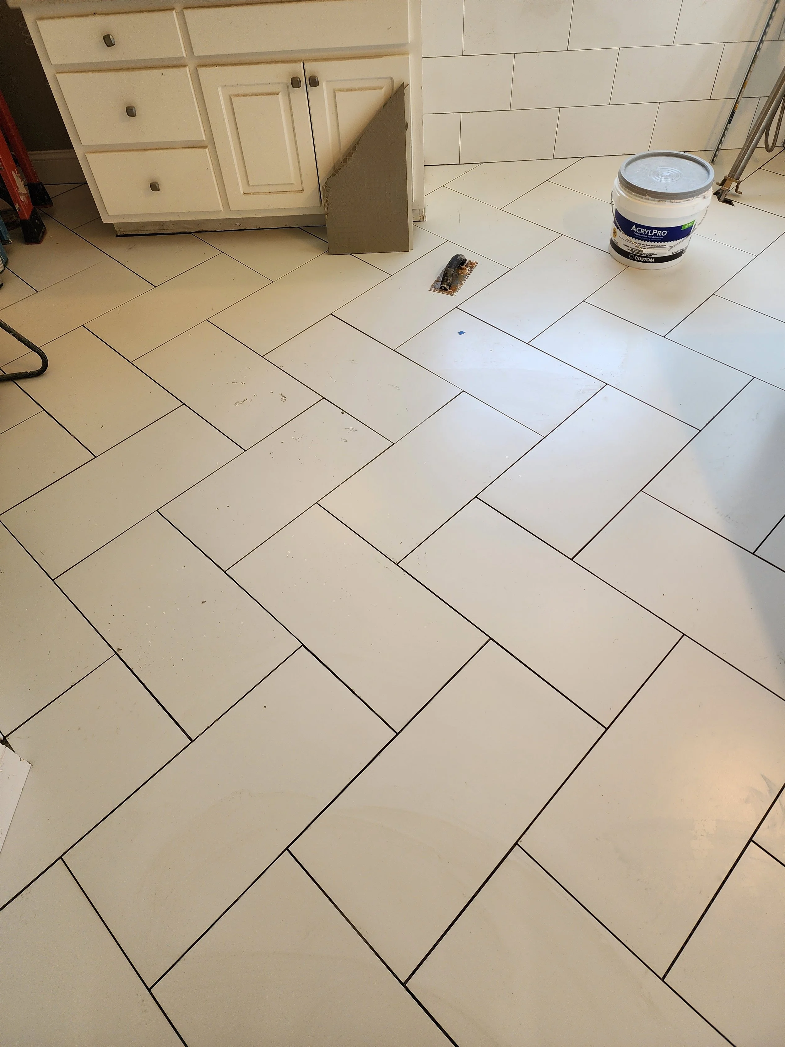 White tile flooring in a kitchen with a bucket of paint, a piece of tile, and a floor scraper.