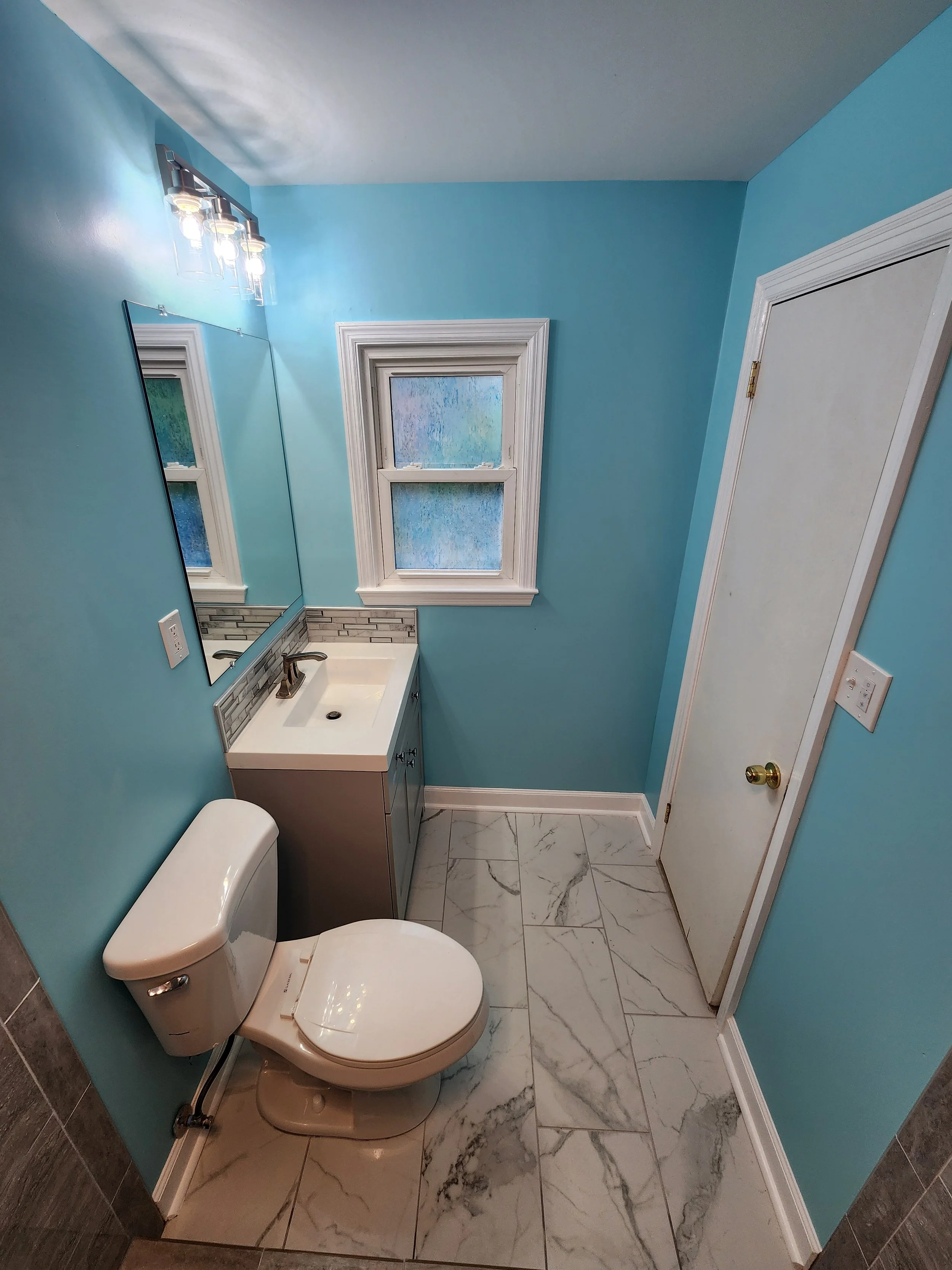 Small bathroom with turquoise walls, white marble floor tiles, a white toilet, a small white vanity with a black faucet, a large mirror, a window with white trim, and a white door.