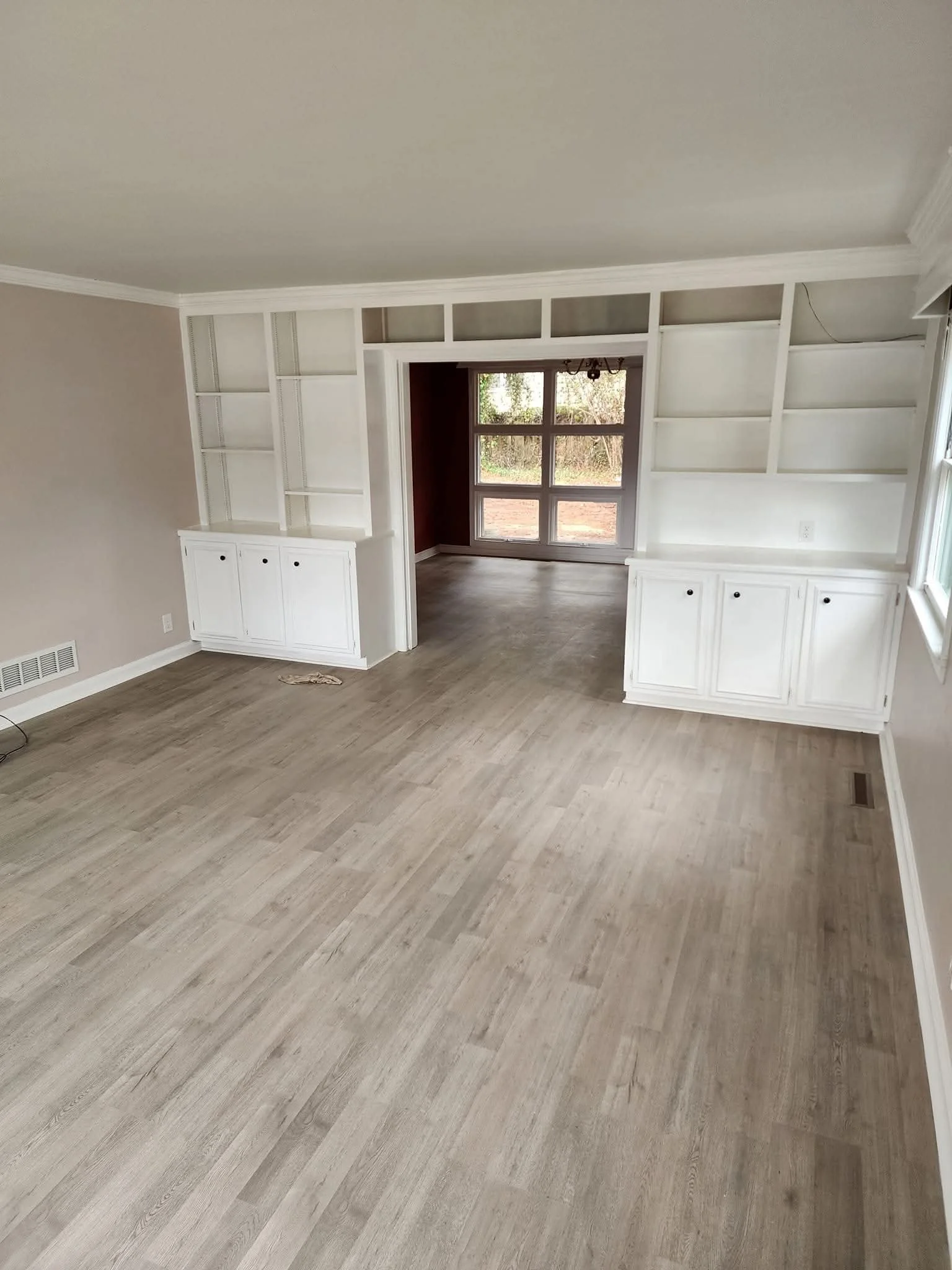 Empty living room with beige walls, light wood flooring, built-in white bookshelves and cabinets, large window, and connecting dining area with a window and glass door leading outside.