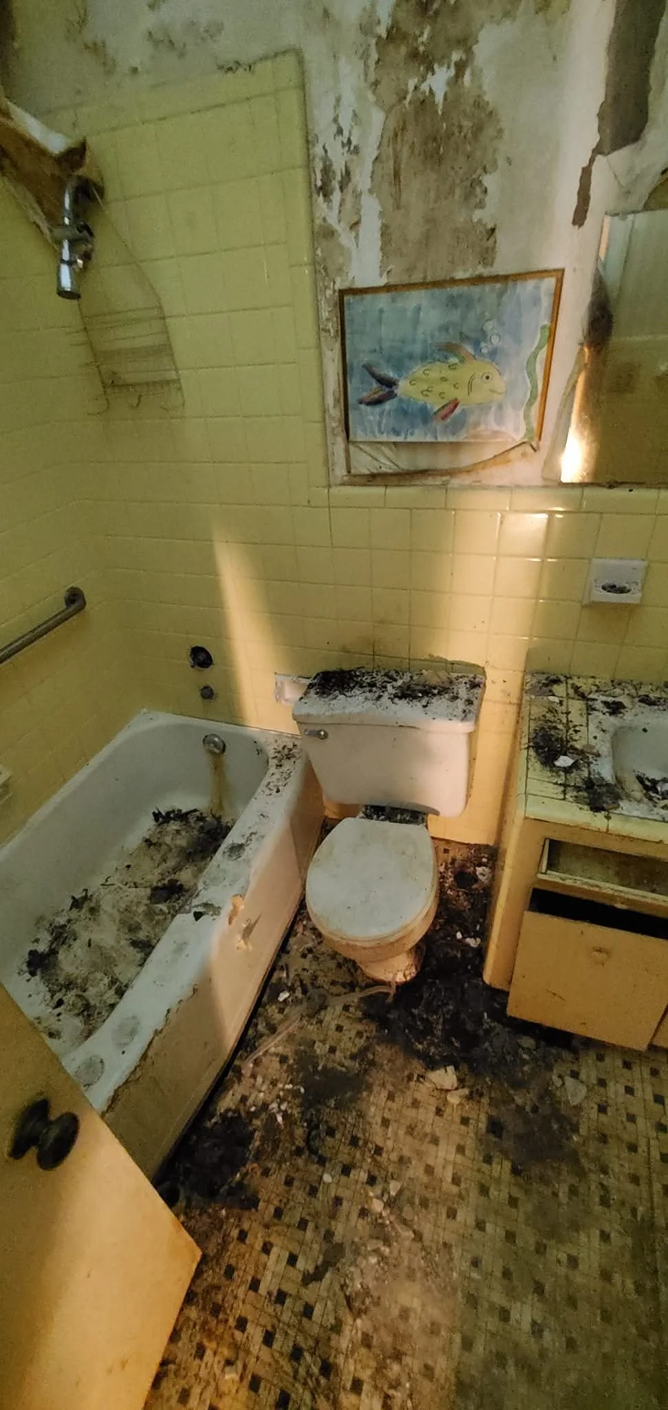 A severely burned and dirty bathroom with a yellow tiled wall, a damaged bathtub, a toilet, a sink, and a damaged mirror. There is a child's fish-themed painting on the wall.