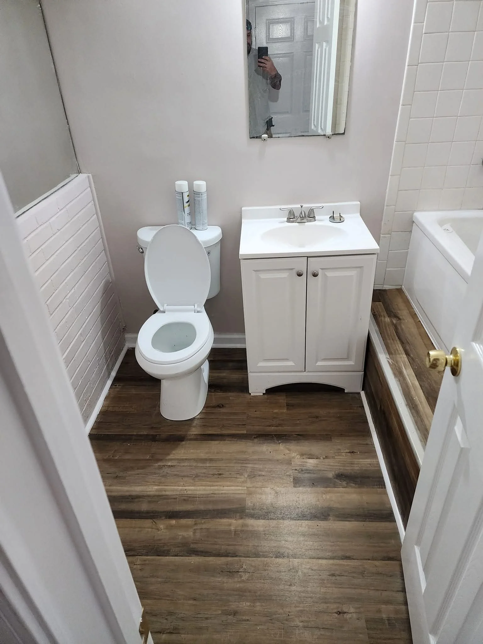 A small bathroom with a toilet, a small white vanity with a sink, a mirror above the sink, a bathtub partially visible on the right, and wood-like flooring.