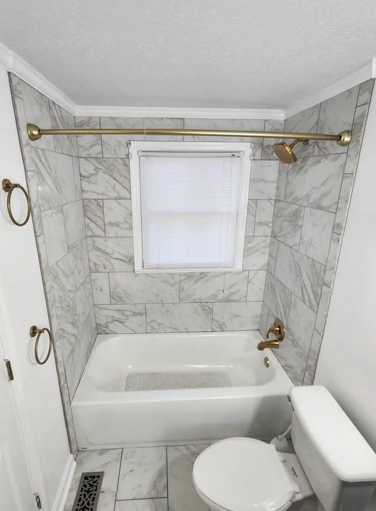 Bathroom with a bathtub surrounded by marble tiles, a window with white blinds above the tub, a window curtain rod, a brass faucet, a brass showerhead, a toilet next to the tub, and two brass towel rings on the wall.