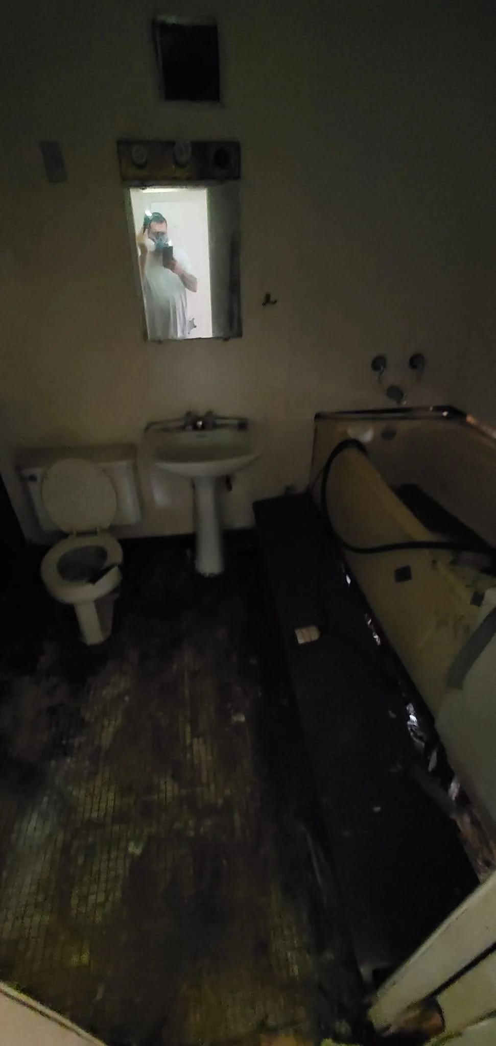 A dark, dirty bathroom with a toilet, a sink, a mirror, and a bathtub. The floor is stained and the bathroom appears abandoned.