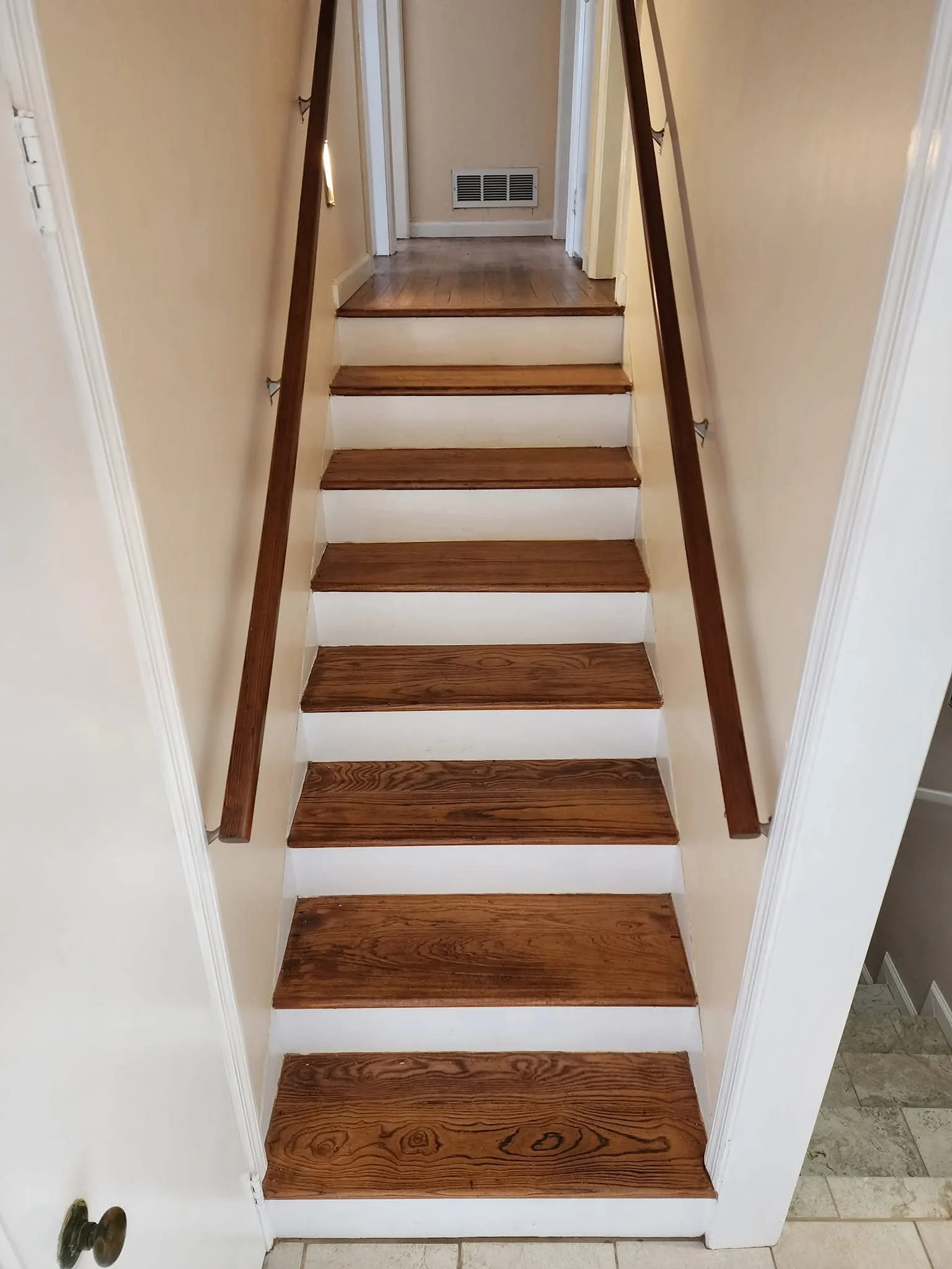 Wooden stairs with brown handrails ascending from tiled floor to a small landing with at least three more steps, leading to a door with a vent on it.