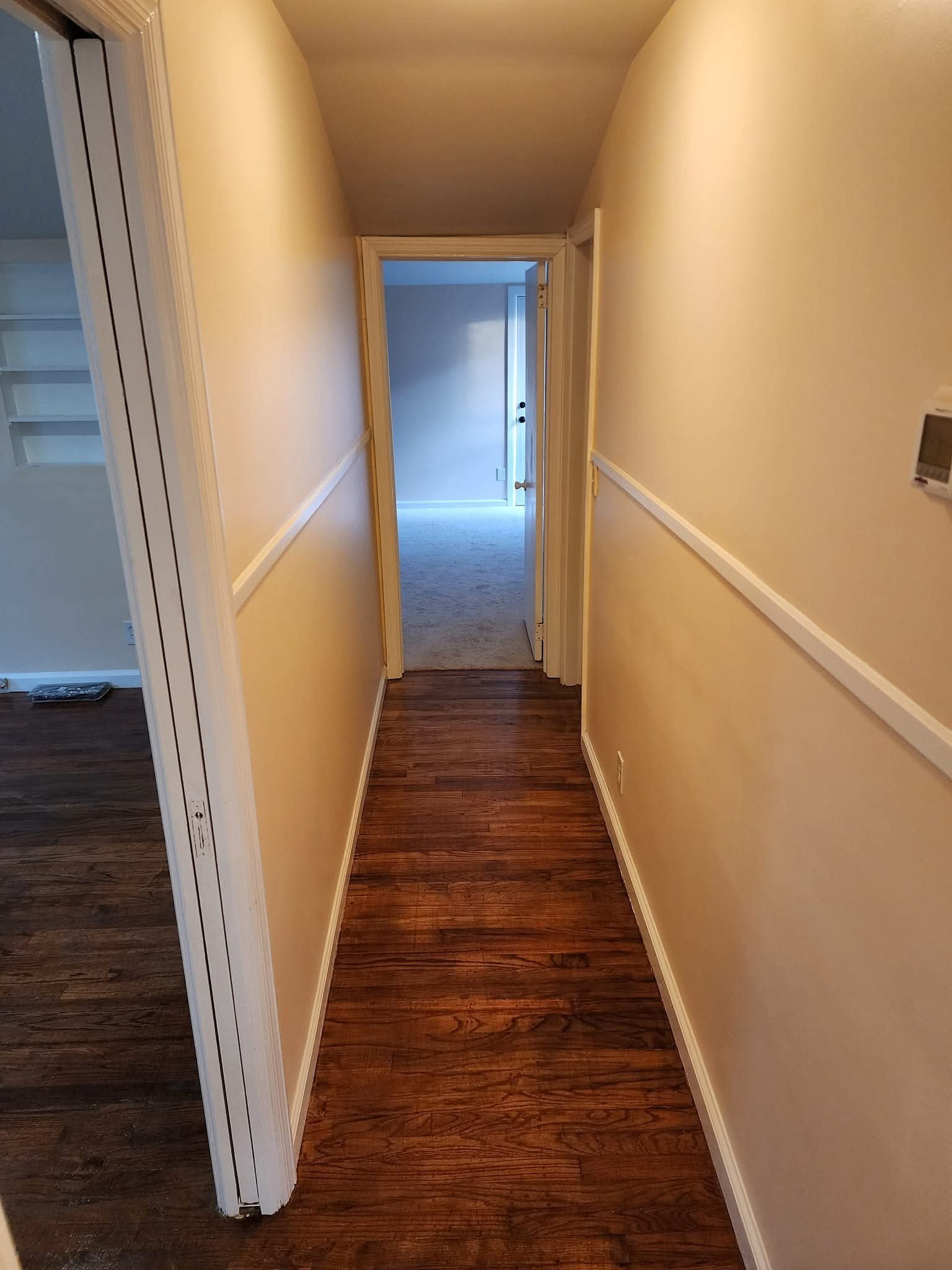 Empty hallway with yellow walls, brown hardwood flooring, and a white door at the end leading to a carpeted room.