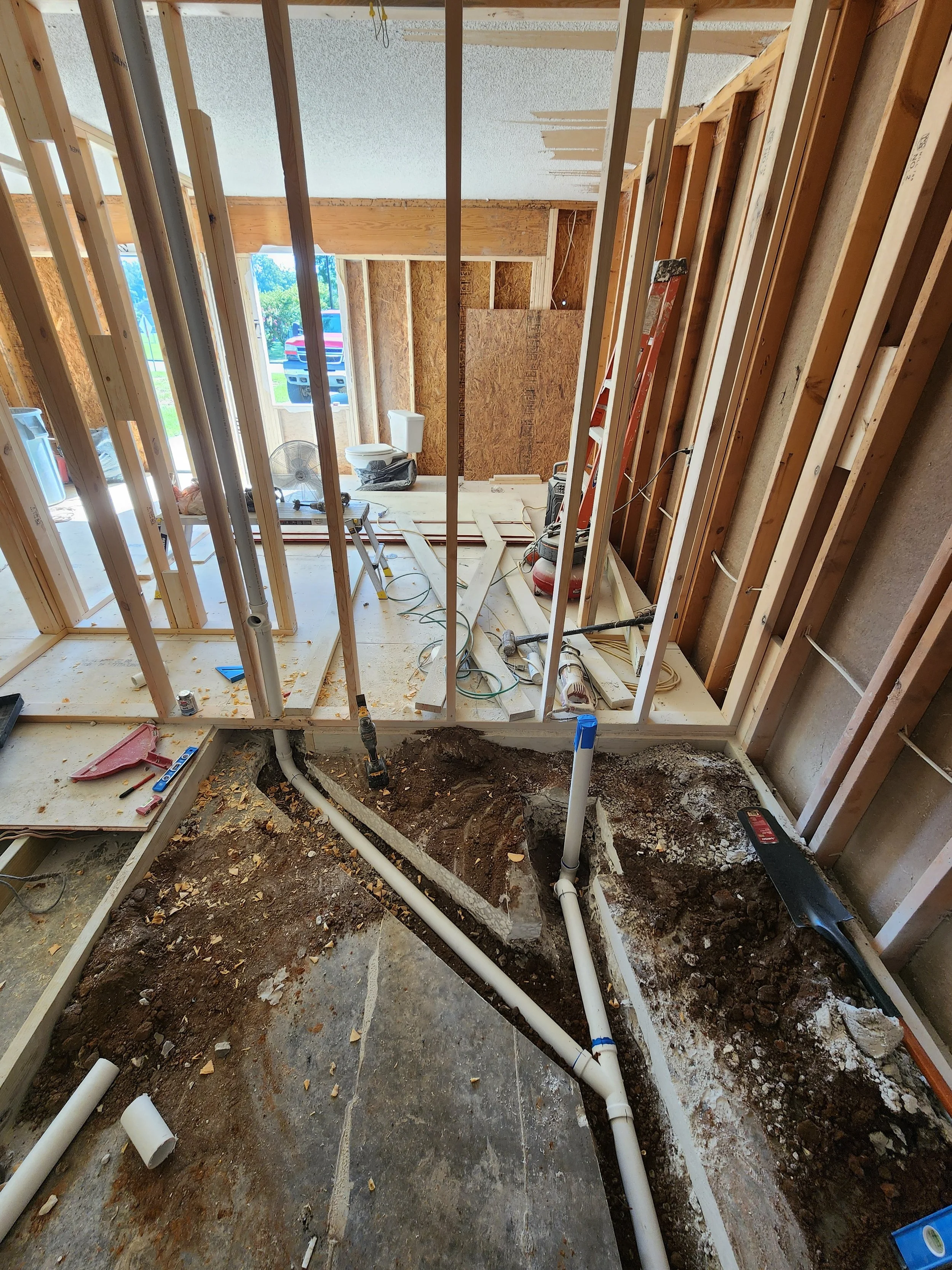 Interior view of a house under construction, showing exposed wooden framing, plumbing pipes, and construction tools on the floor.