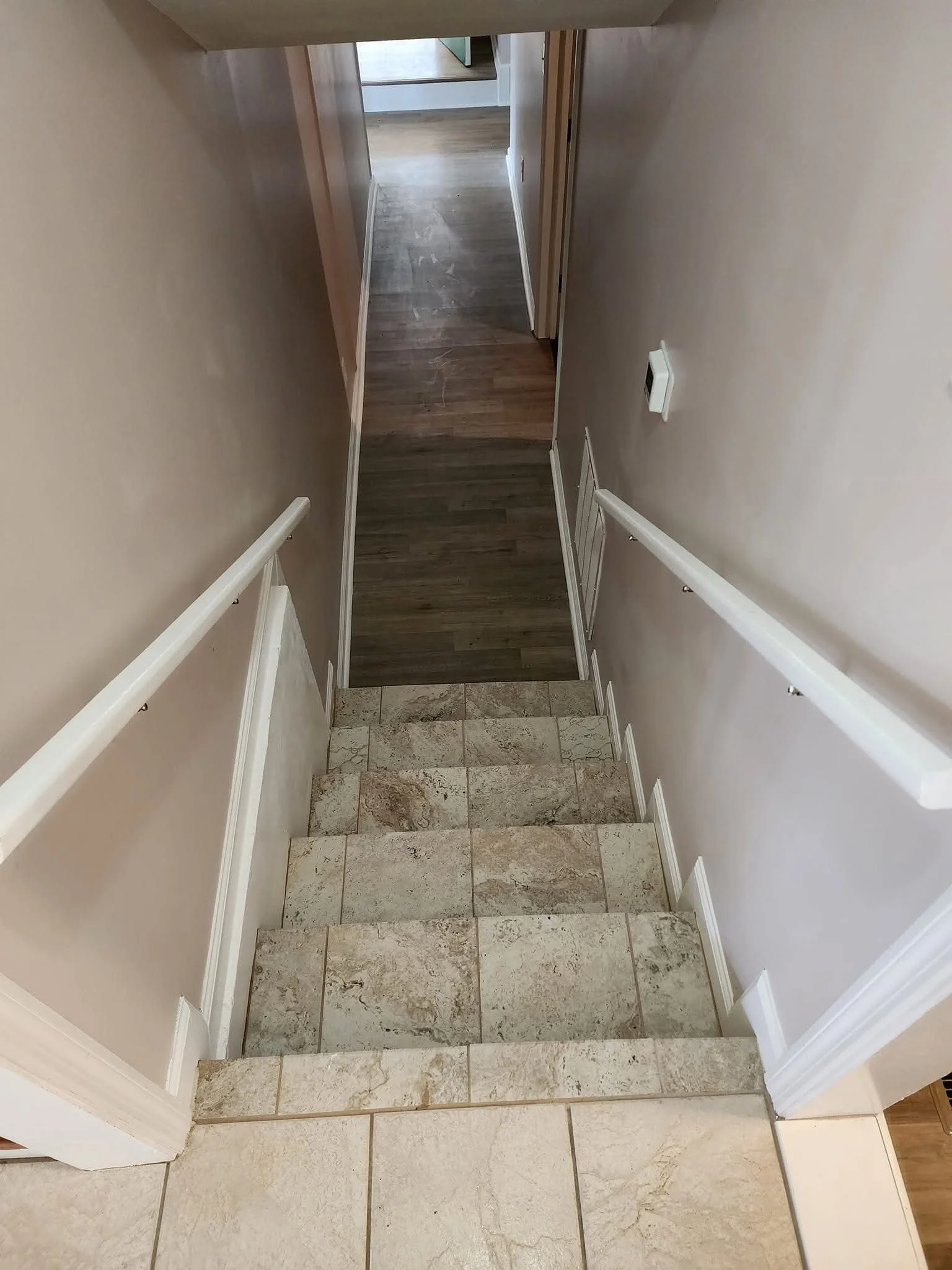 View of a staircase with beige tiles, white handrails, and a hallway with hardwood flooring leading to other rooms.