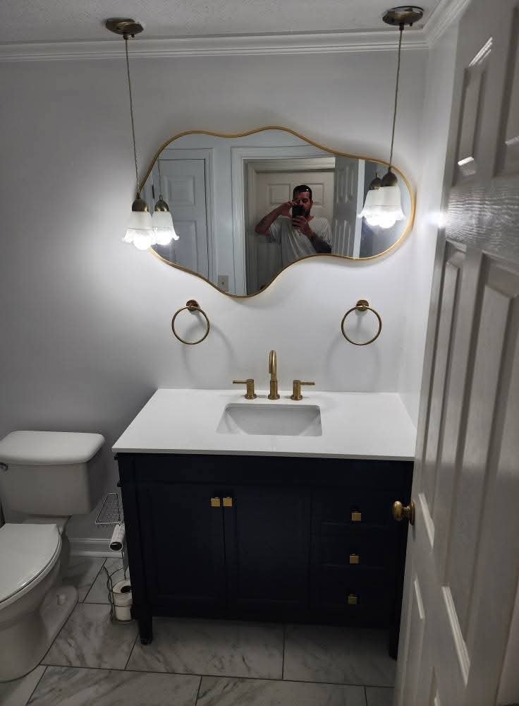 A bathroom with a black vanity, white countertop, gold fixtures, and a uniquely shaped mirror. Two pendant lights hang on either side of the mirror, and two gold towel rings are mounted on the wall. A toilet is visible to the left, and the floor is t