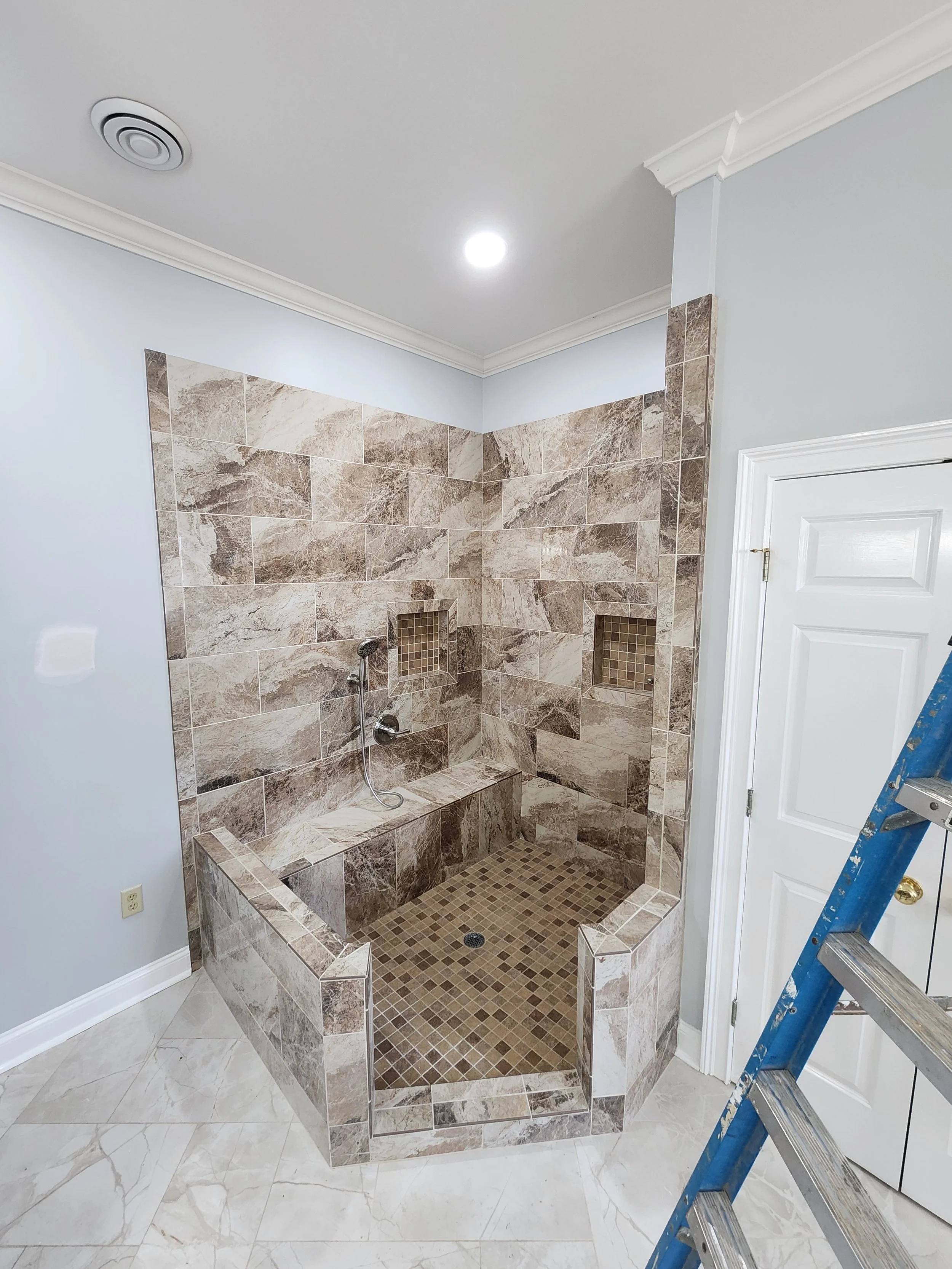 A bathroom shower area with tiled walls and floor, featuring two built-in shelves, a handheld shower head, and a small drain on the floor. There is also a white door and a blue ladder in the scene.