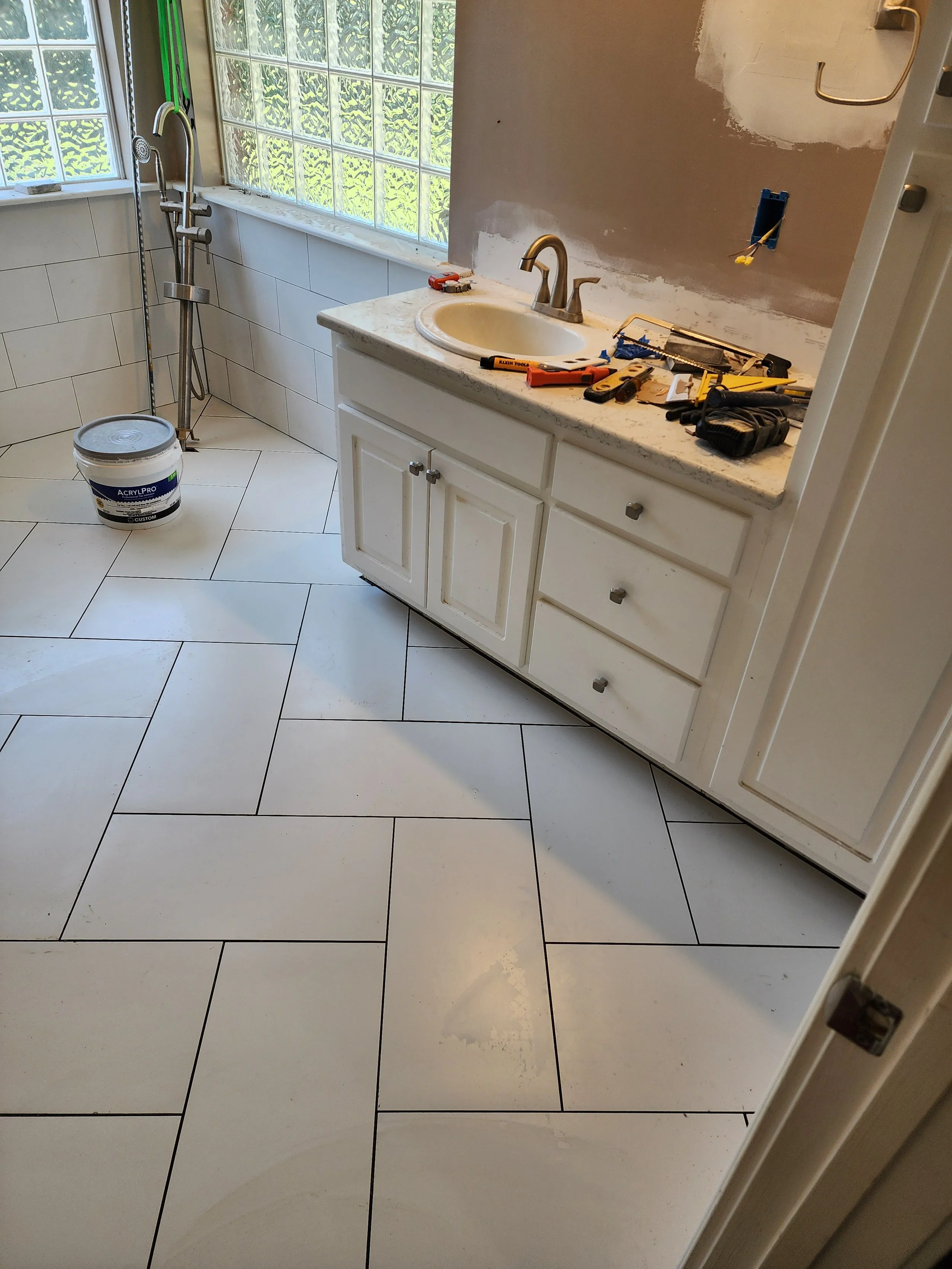 A bathroom under renovation with a white tile floor, a white vanity with a sink, yellow and black tools on the countertop, a bucket, a partially painted wall, and large windows with green foliage outside.
