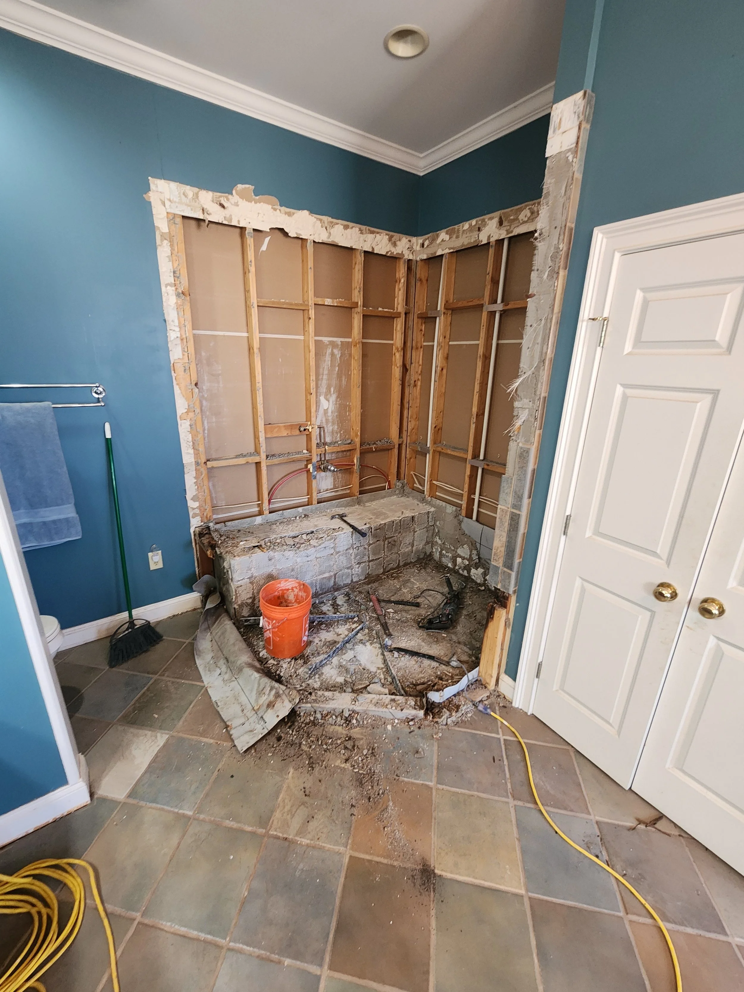 Bathroom with a wall removed, exposing wall studs and a brick foundation. Construction tools and materials, including a bucket, hammer, and power drill, are on the floor. The room has colorful tile flooring and a white door.