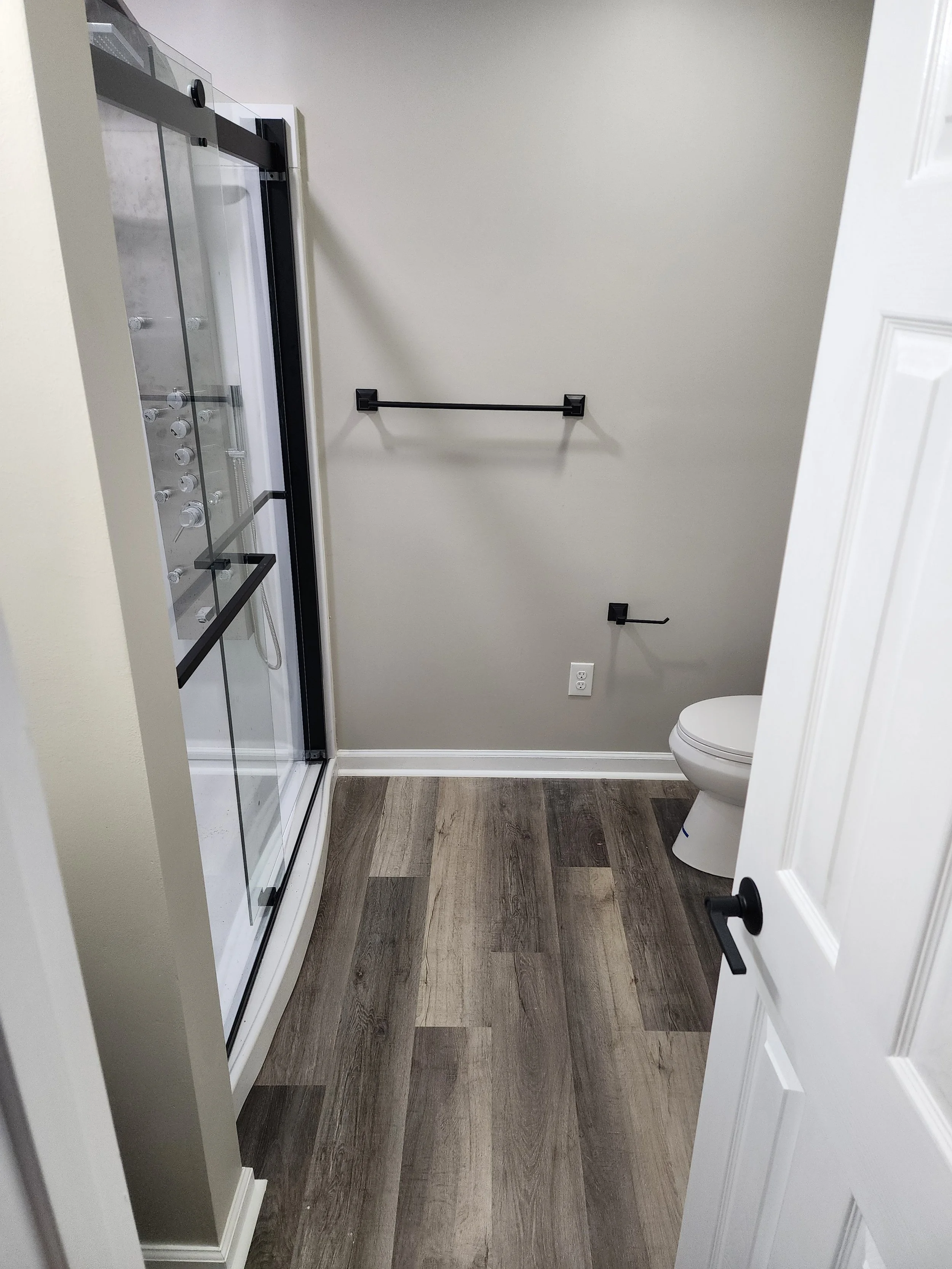 Empty bathroom with a shower stall, a toilet, and wall-mounted black fixtures, including a towel rack and toilet paper holder, with wood-look flooring and a wall outlet.