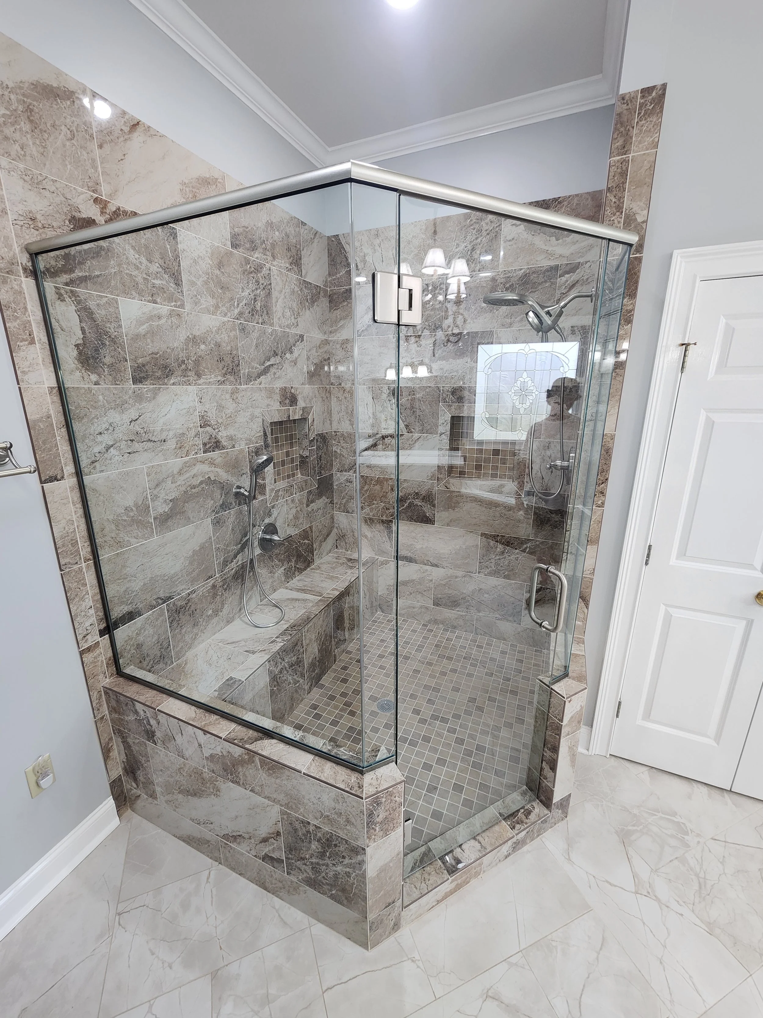 A glass shower enclosure with natural stone tiles, built-in bench, and two shower heads, set in a bathroom with marble flooring and white door.