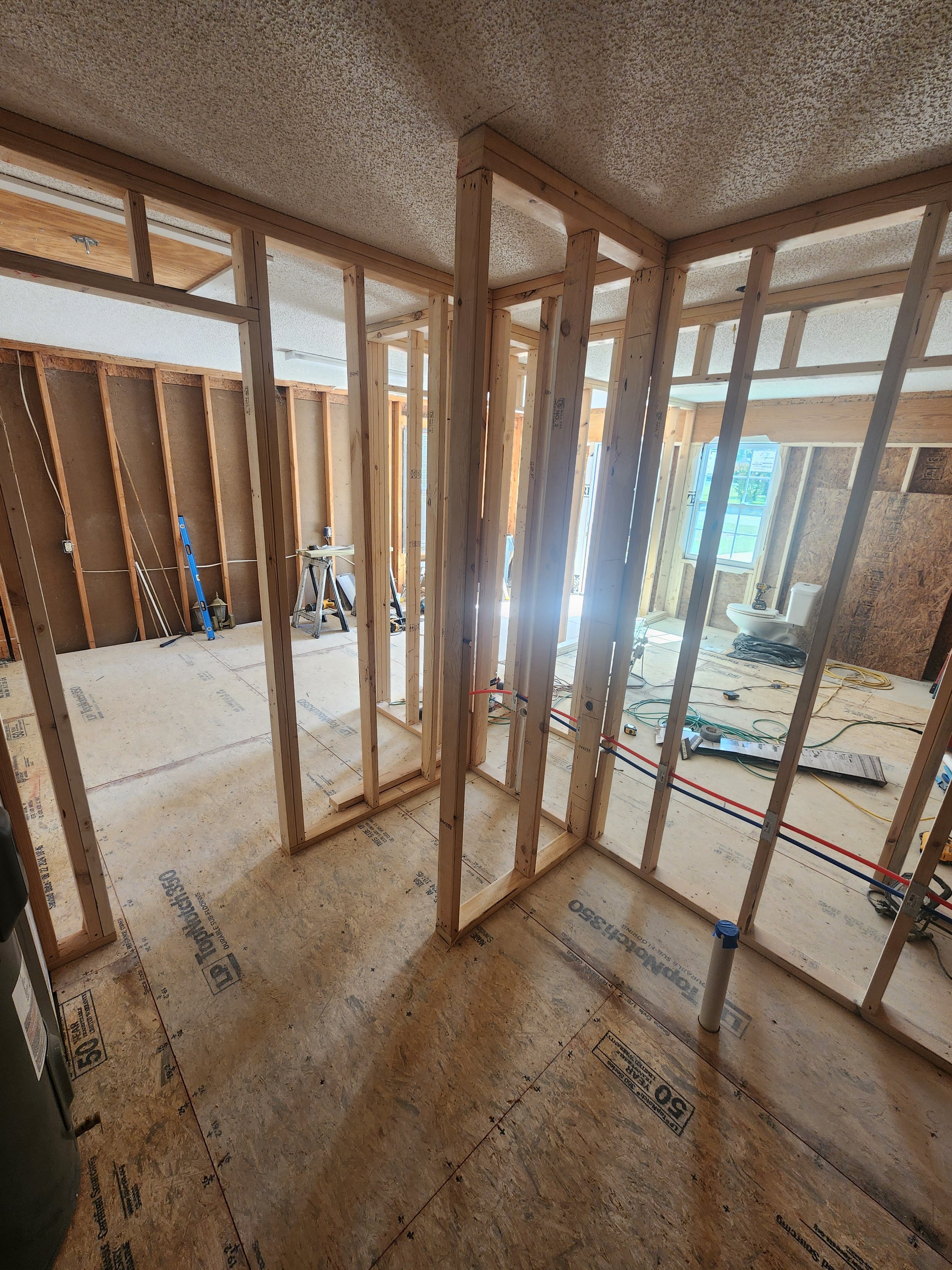 Interior view of a house under construction with visible wooden framing, electrical wiring, and plumbing pipes.