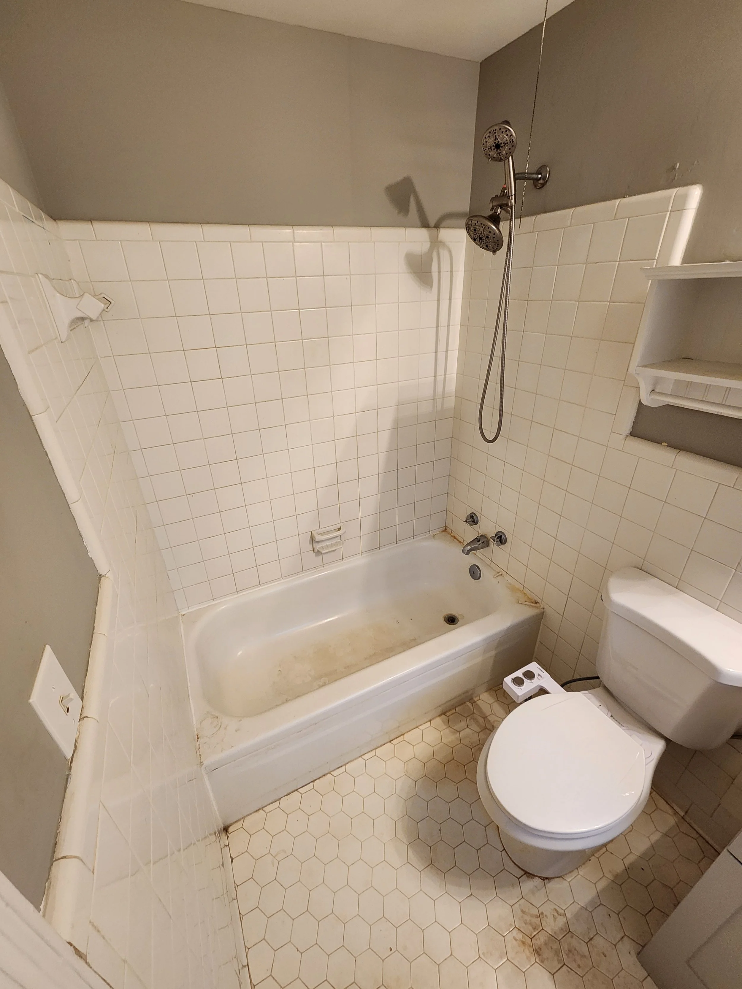 A bathroom with a bathtub, toilet, and shower. The bathtub has stained, dirty corners. The walls have white tiles, and the floor has hexagonal tiles with visible dirt. The showerhead is mounted on the wall, and there is a small shelf above the toilet