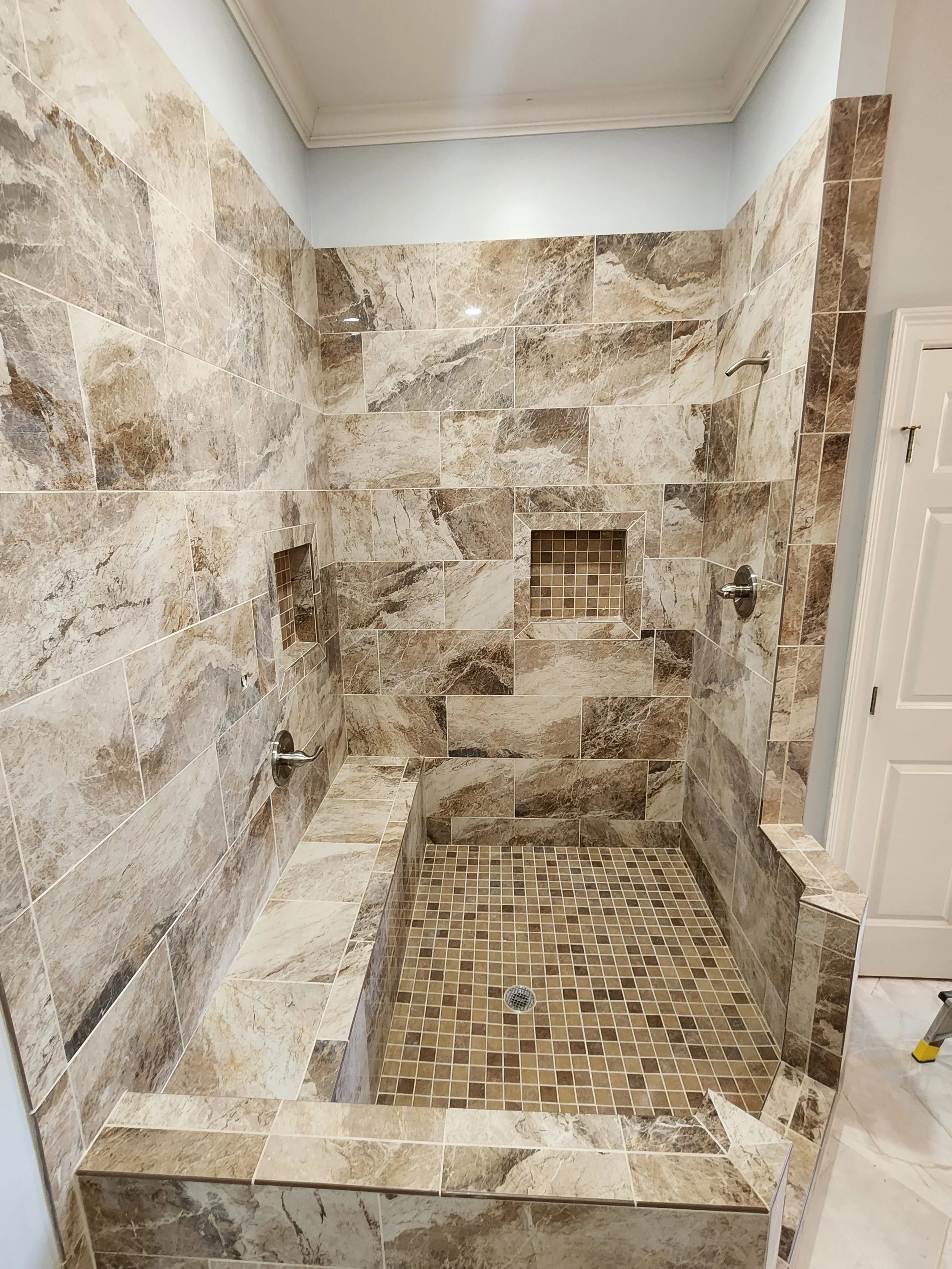 A walk-in shower with beige and brown marble tiles on the walls and small square brown mosaic tiles on the shower floor and inside two wall niches, with metal shower fixtures and a built-in bench.