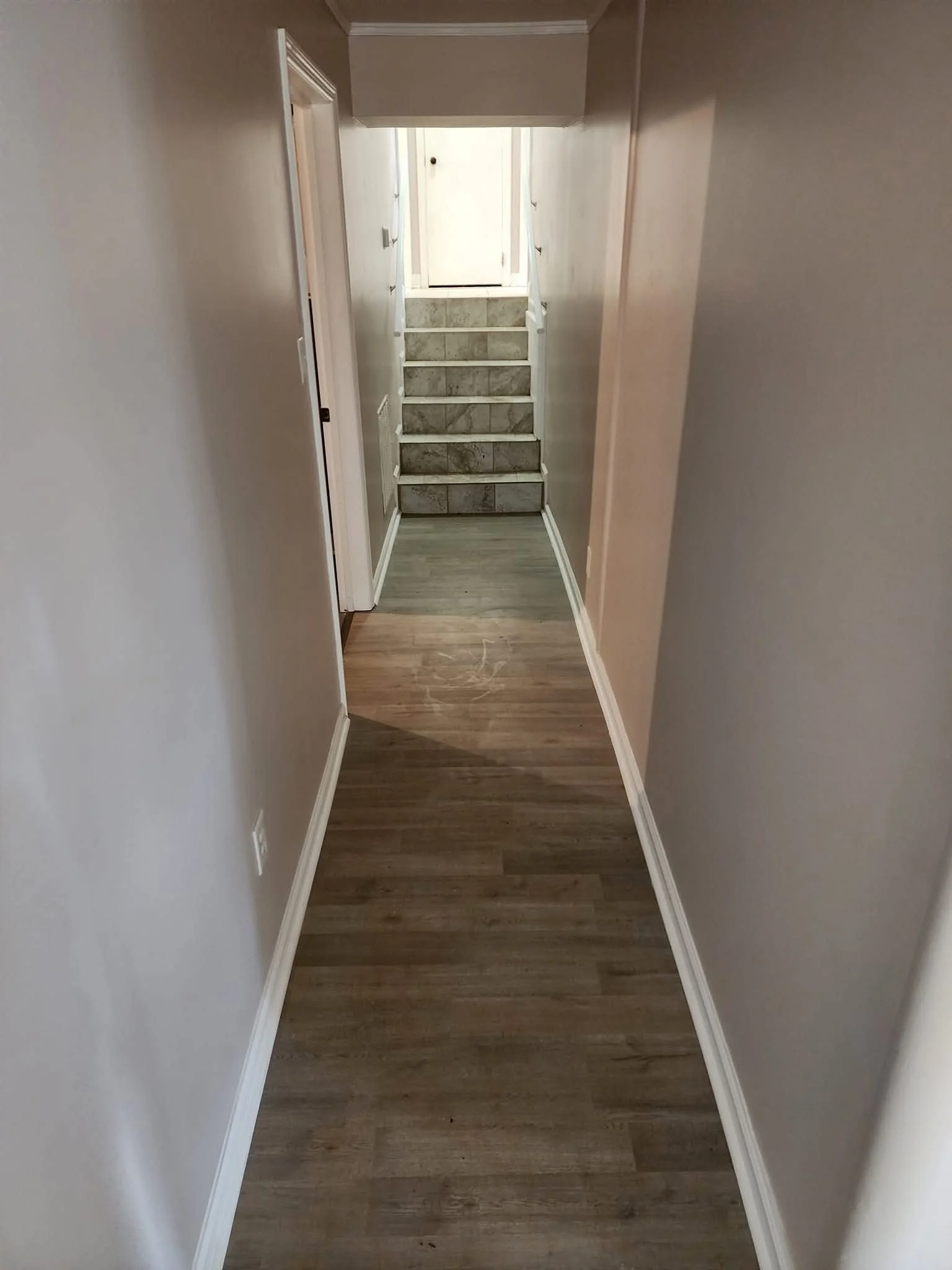 A narrow hallway with light-colored walls and wooden flooring leading to a carpeted staircase at the end, with a door at the top of the stairs.
