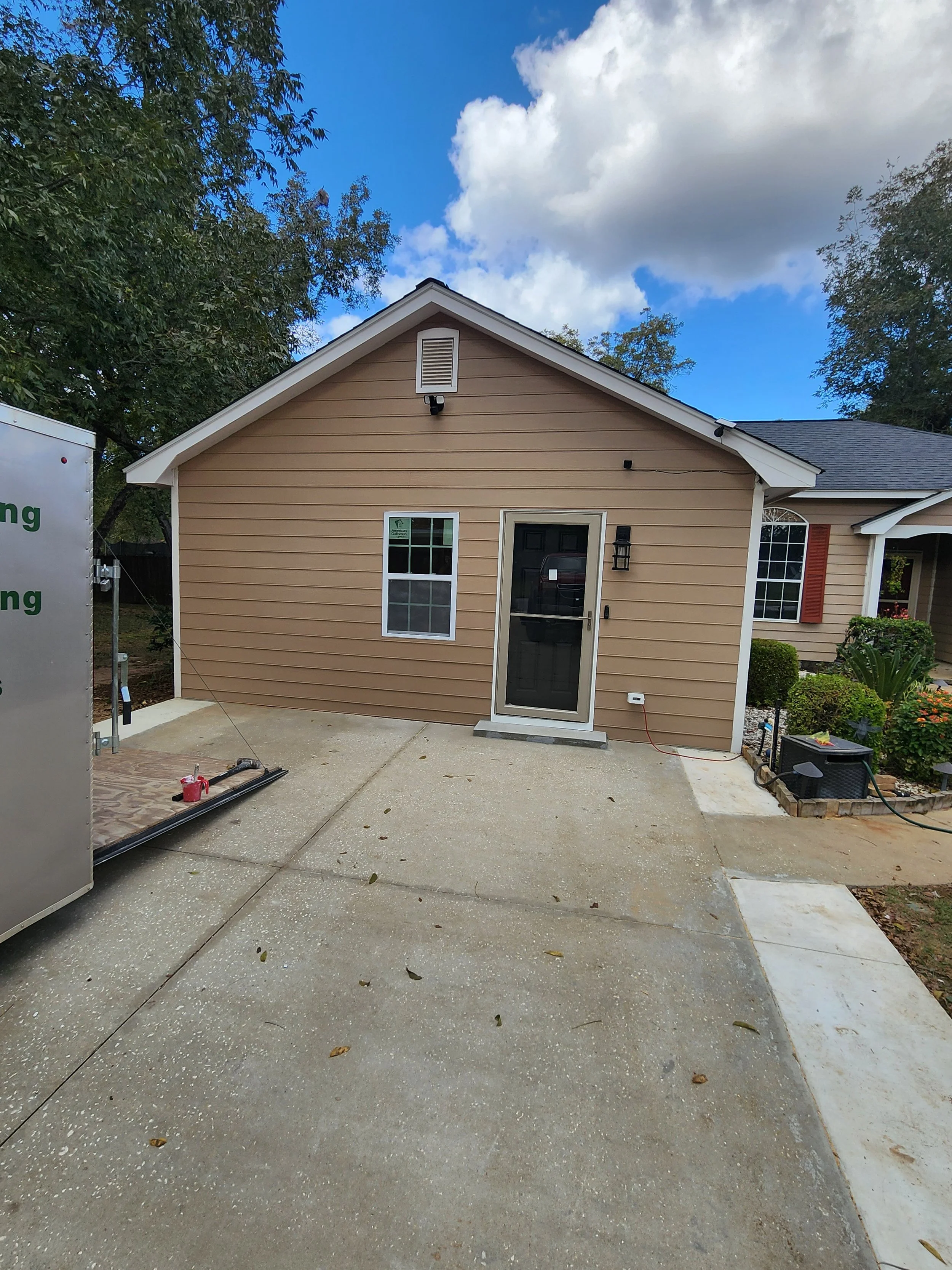 A newly constructed beige house with white trim, a black front door with a screen door, and a small concrete step. There is a window next to the door, outside black lantern-style light fixtures, and a small front garden with shrubs and plants. The co