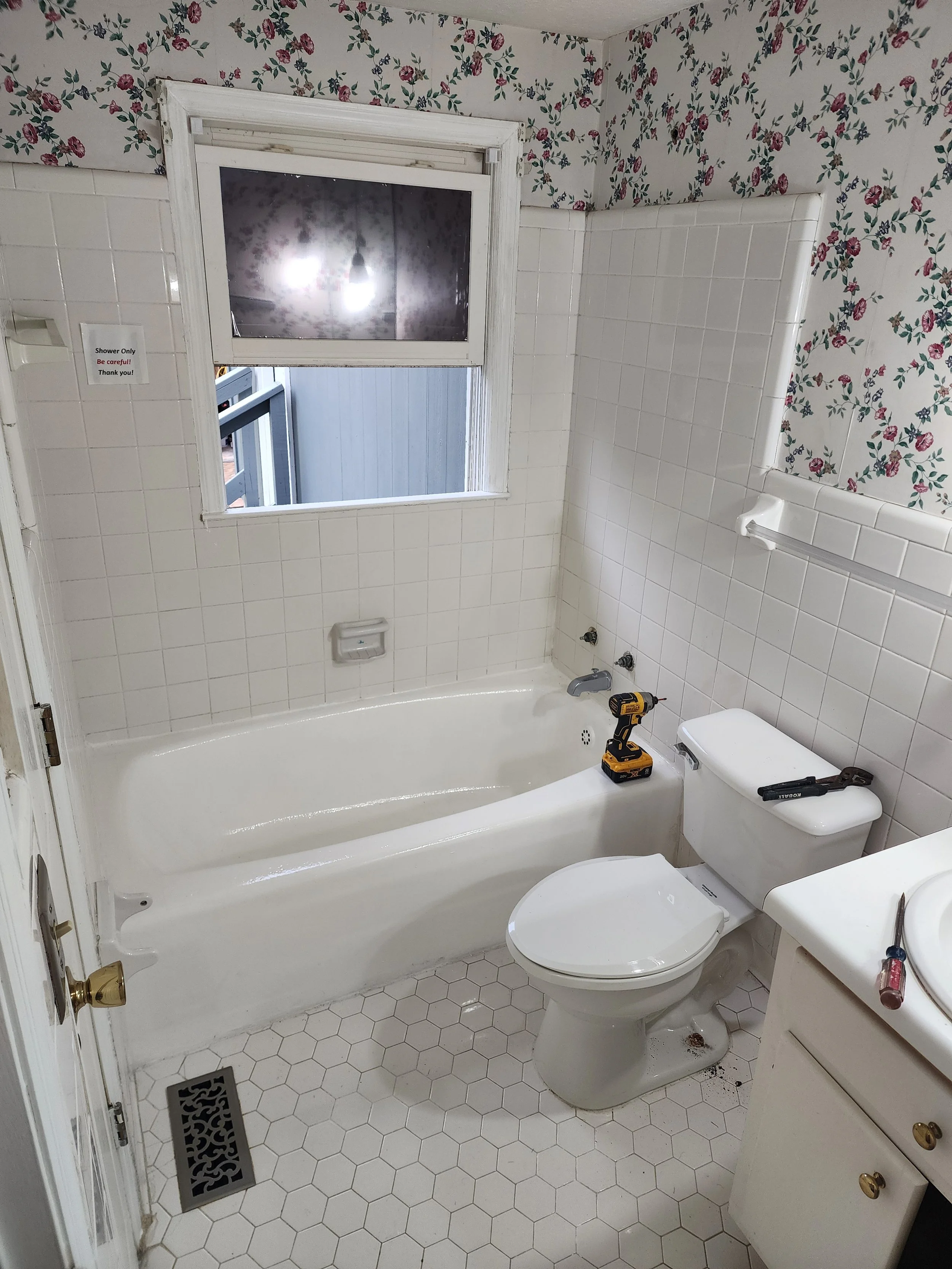 Small bathroom with floral wallpaper, white tiles, and hexagonal floor tiles. The bathtub is under a window with wood trim, and a yellow drill is on the toilet tank. The bathroom has a small sink with a screwdriver on the counter.