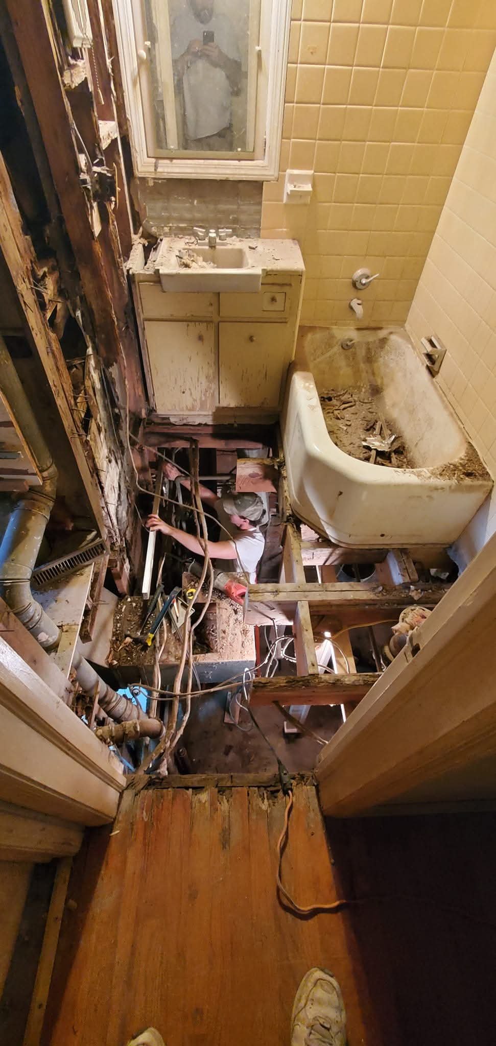 A bathroom under renovation showing a partially removed wall, exposed pipes, a dirty bathtub, a sink with a mirror, and a person working in the space.