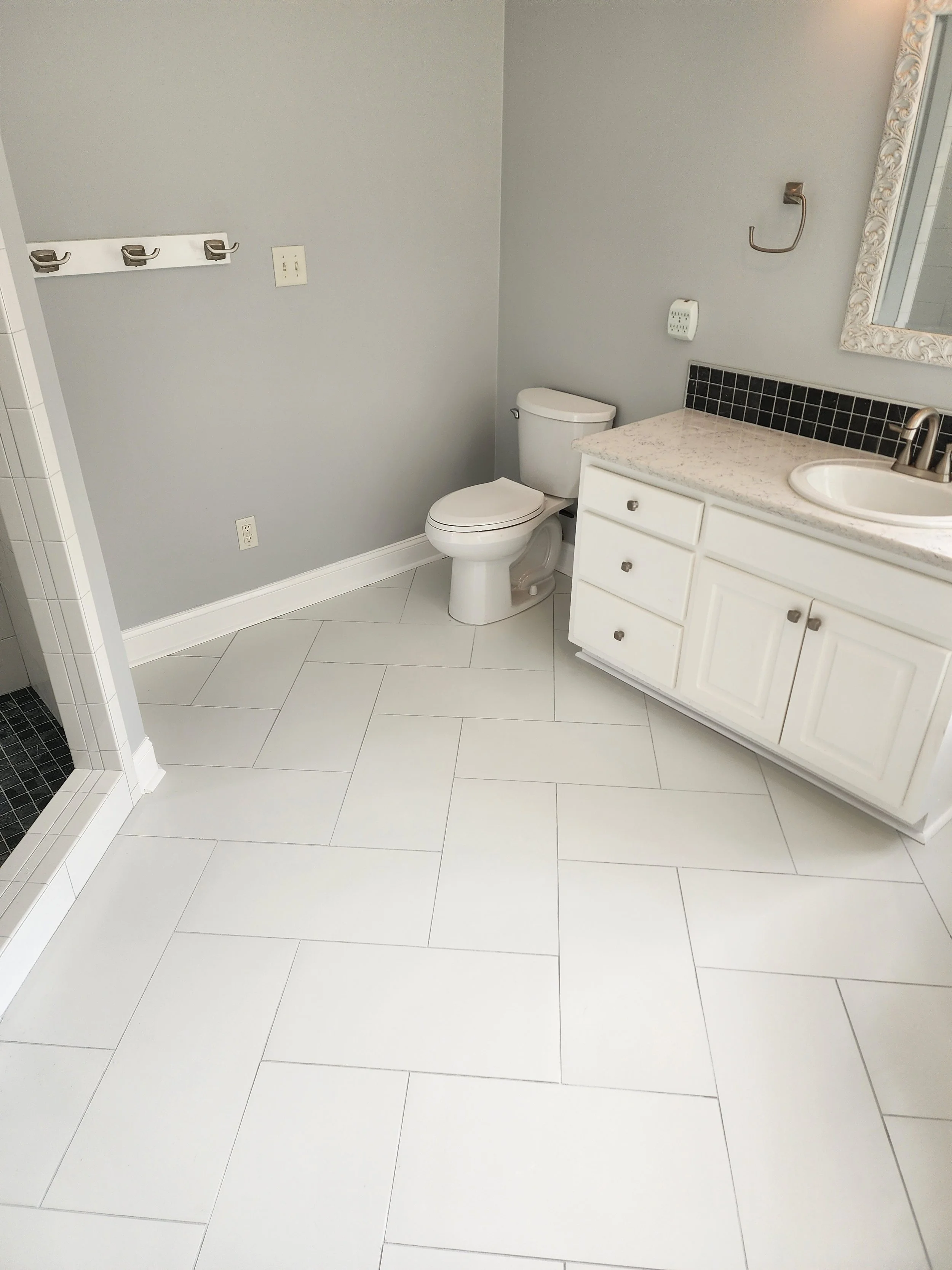 Empty bathroom with white tiled floor, light gray walls, a toilet, a white vanity with a marble countertop and a sink, a mirror, and some wall hooks.