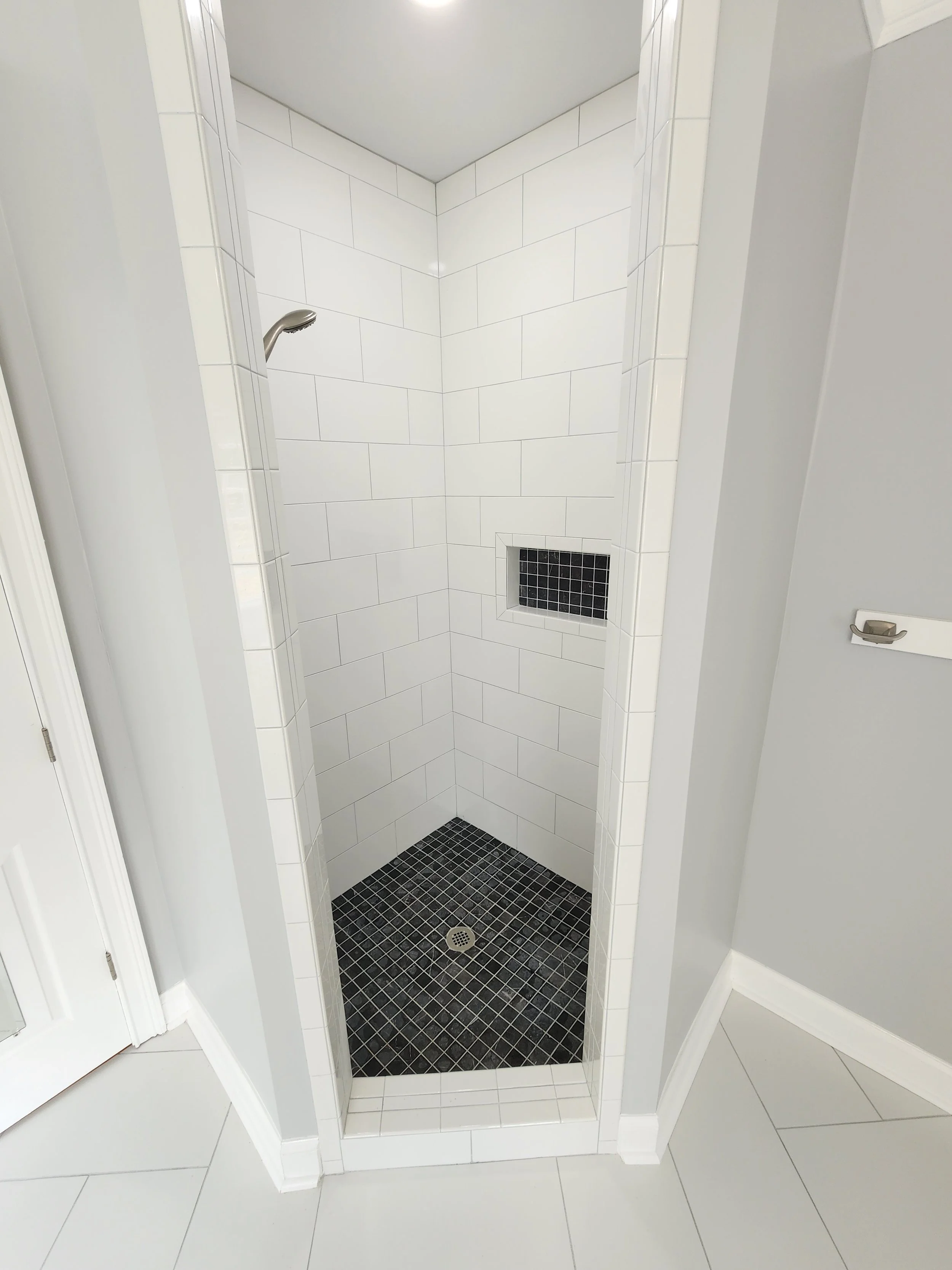 Small corner shower with white tiled walls, black small square floor tiles, a wall niche with black tiles, and a silver showerhead.