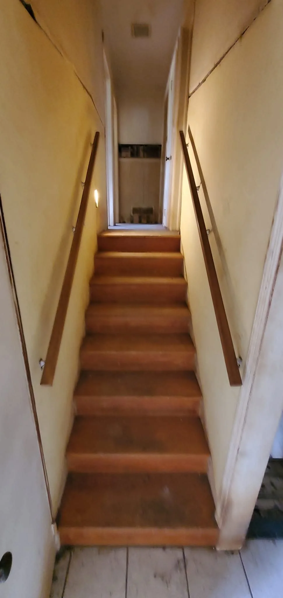Photo of a small, steep wooden staircase with handrails on both sides leading to a closed door at the top, in a narrow hallway with yellow walls.