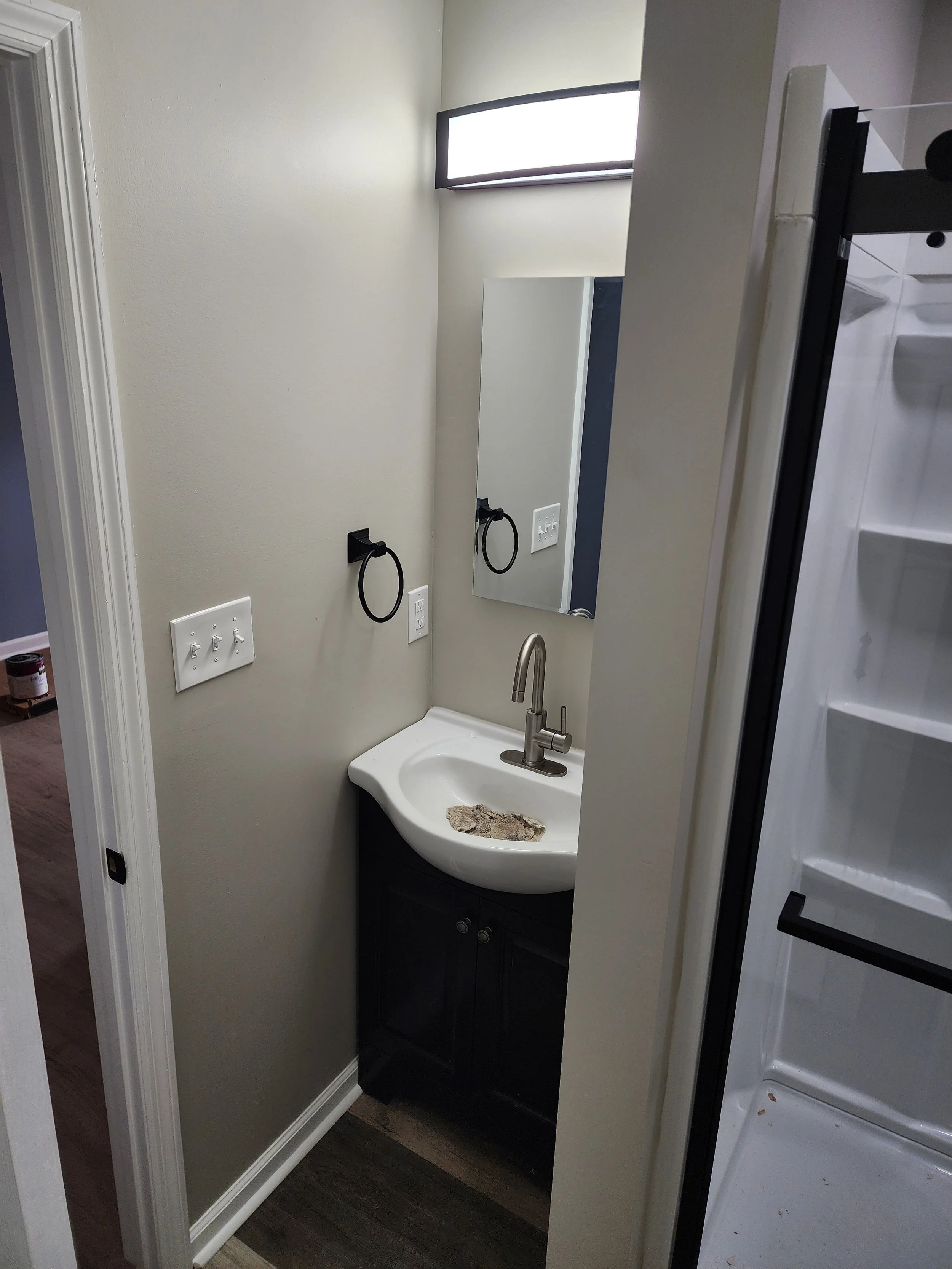 Small bathroom with a white sink, dark cabinet, small mirror, black towel ring, and gray wall with a light fixture above.