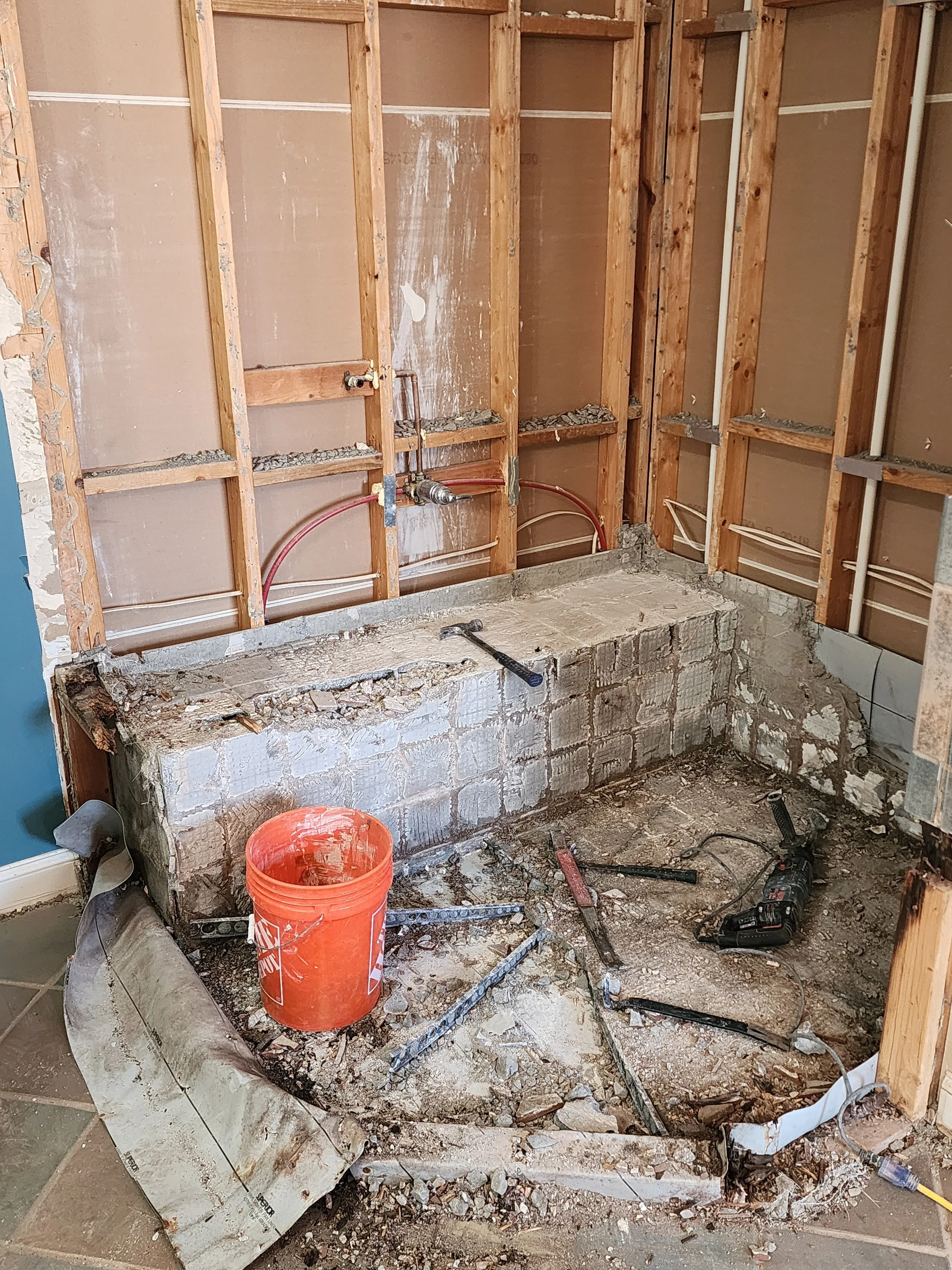 Construction site with exposed wall studs, a partially demolished concrete structure, tools, a bucket, and debris on the floor.