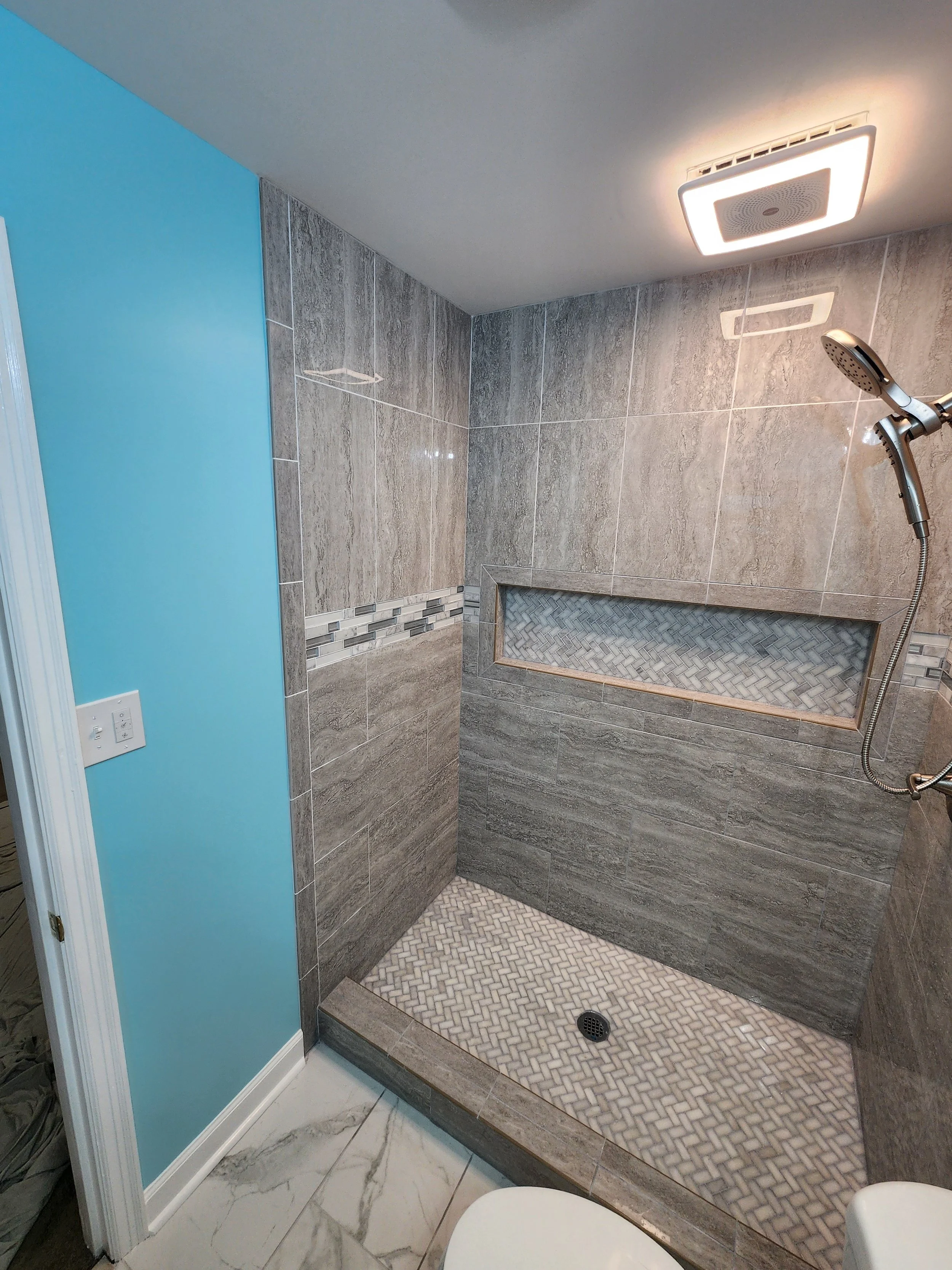 Modern shower area with gray tile walls, a built-in niche with herringbone tile, a rainfall showerhead, and a handheld showerhead. The floor is made of mosaic tiles, and there is a ceiling light.