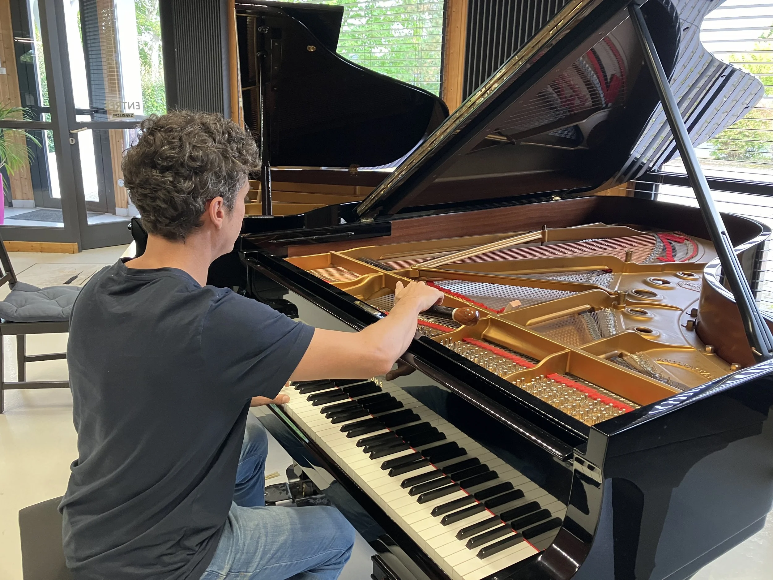 A person with curly hair playing a grand piano indoors, with sunlight coming through large windows and a door in the background.