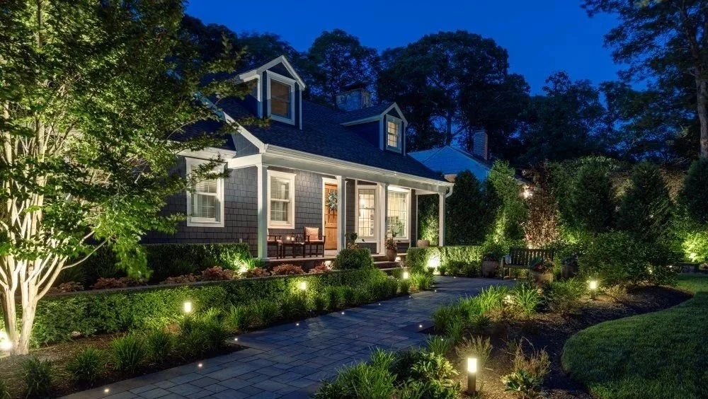 A house with a front porch illuminated at night, surrounded by a landscaped garden with path lighting, trees, bushes, and a bench.