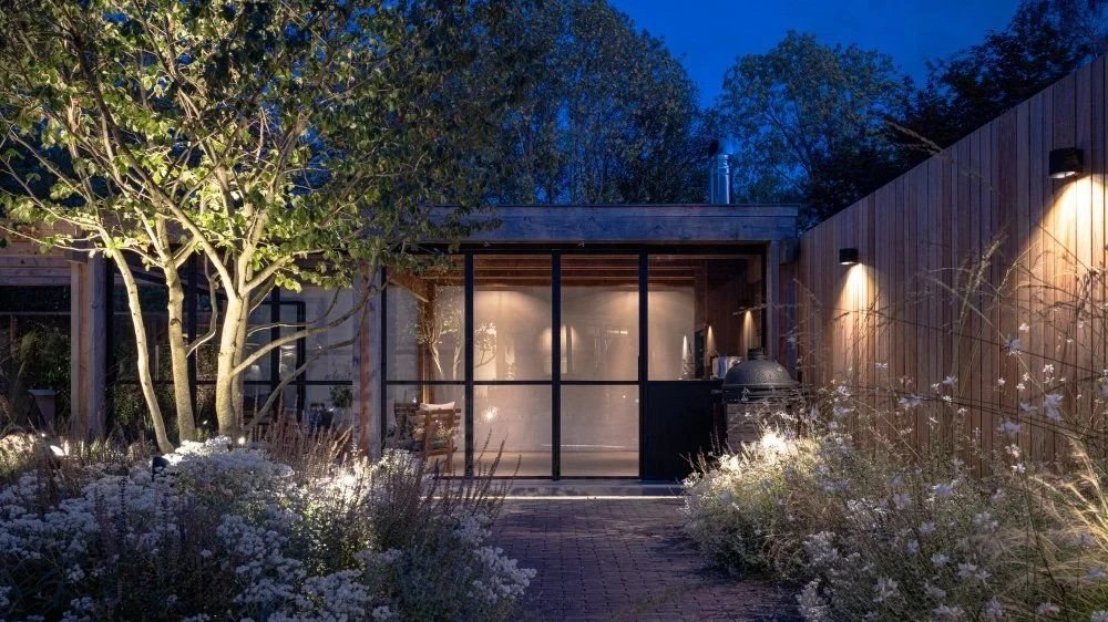 Nighttime view of backyard with illuminated trees and bushes, modern wooden fence, and a glass-sided building or room with outdoor kitchen area.