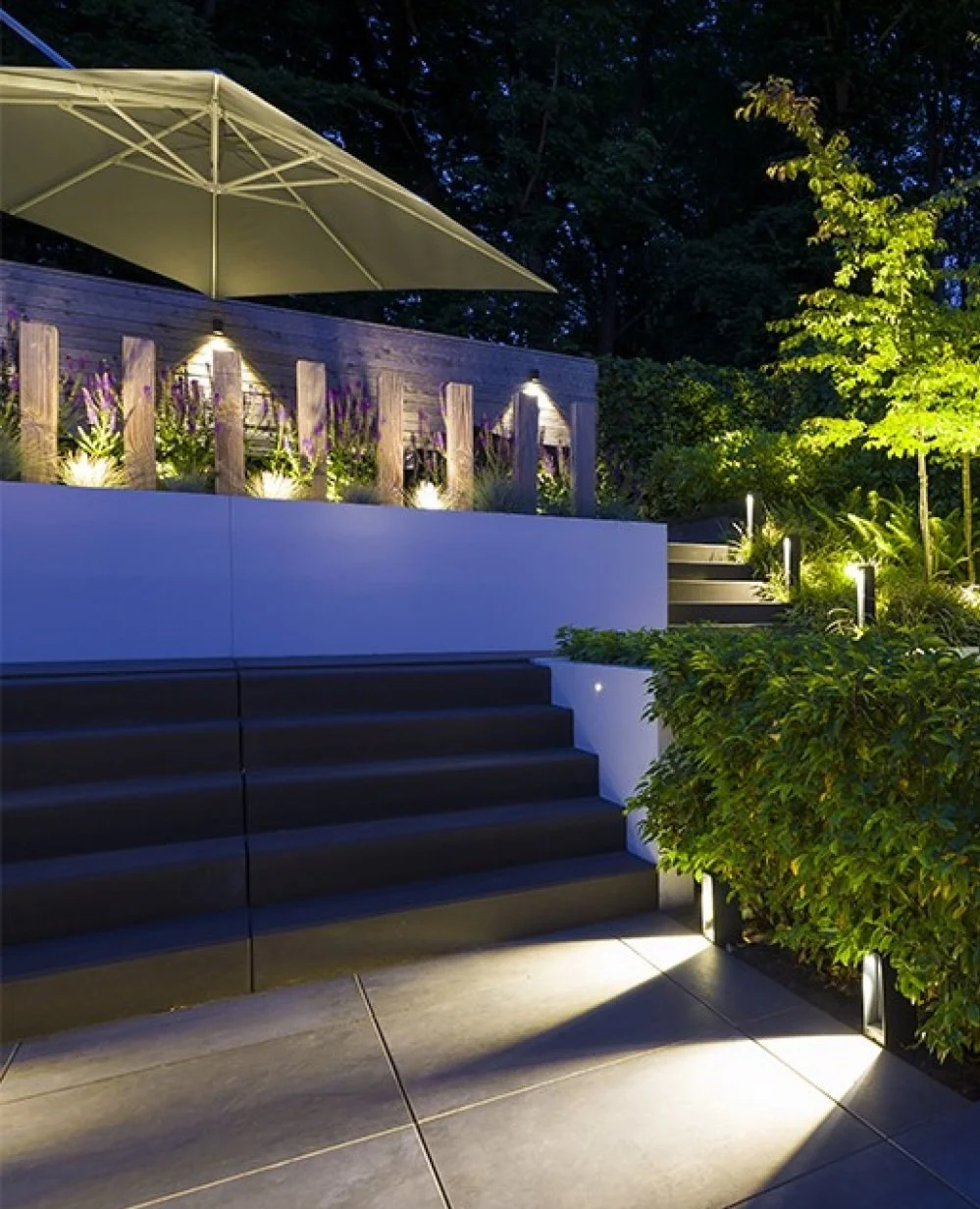 Night view of an outdoor patio with steps, illuminated bushes, and garden lights, featuring a large patio umbrella and wooden fence with purple flowers