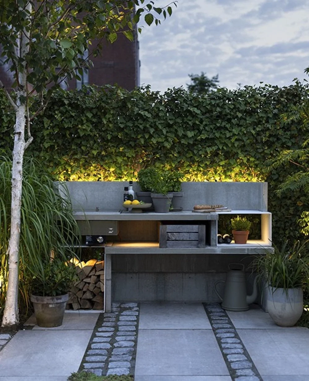 Outdoor kitchen with concrete countertops, potted plants, and a built-in grill, set against a green hedge and evening sky.