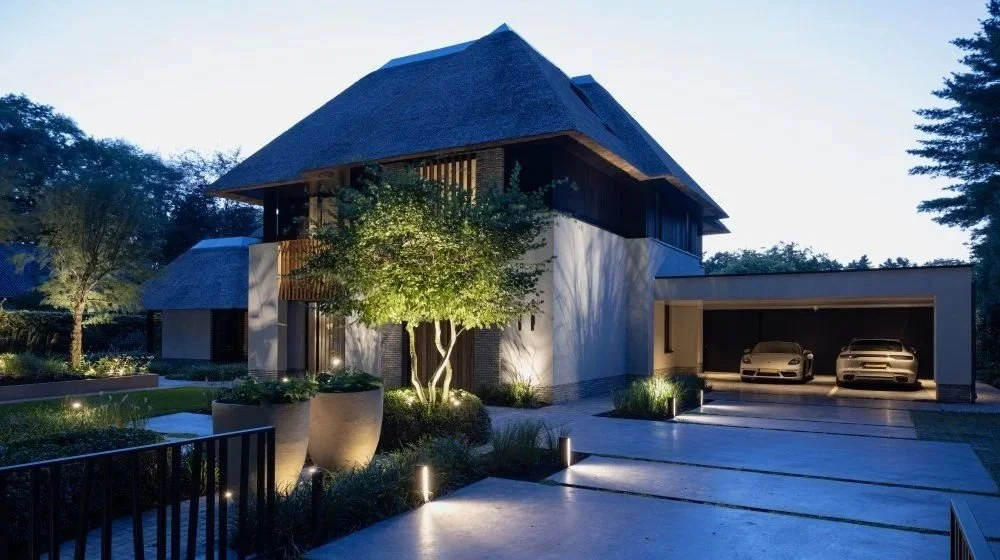 A modern two-story house with a thatched roof, surrounded by landscaped garden with lit pathway and trees, and a garage with two cars inside.
