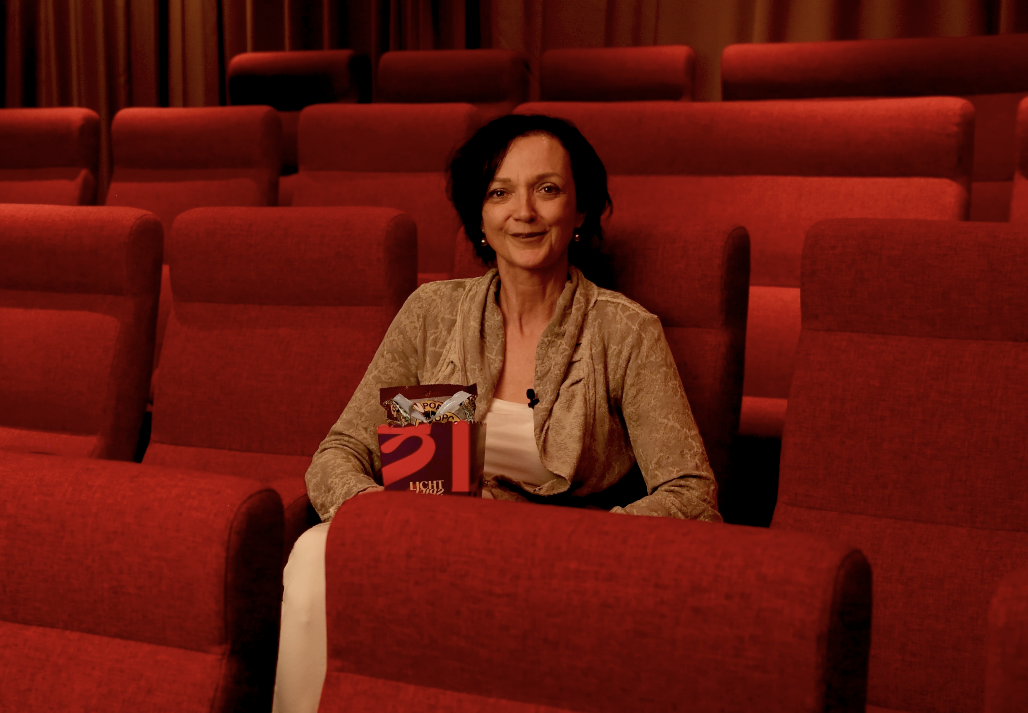 Eine Frau sitzt in einem Kino mit roten Sitzen, hält eine Tüte Snacks in der Hand.