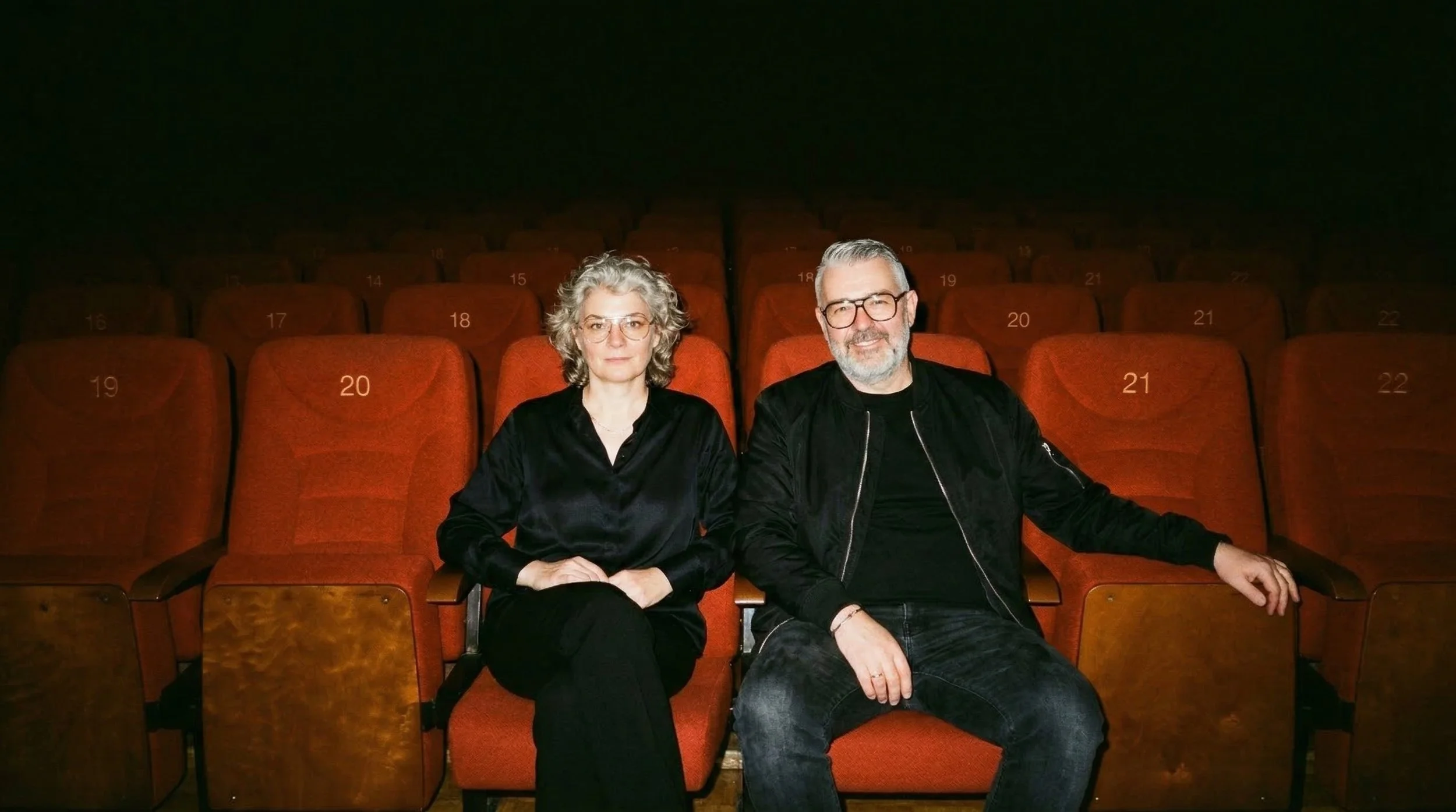 Ein Mann und eine Frau sitzen in einem Kino, umgeben von roten Sitzen mit Nummern. Der Mann trägt eine schwarze Jacke und Brille, die Frau trägt eine schwarze Bluse und auch Brille.