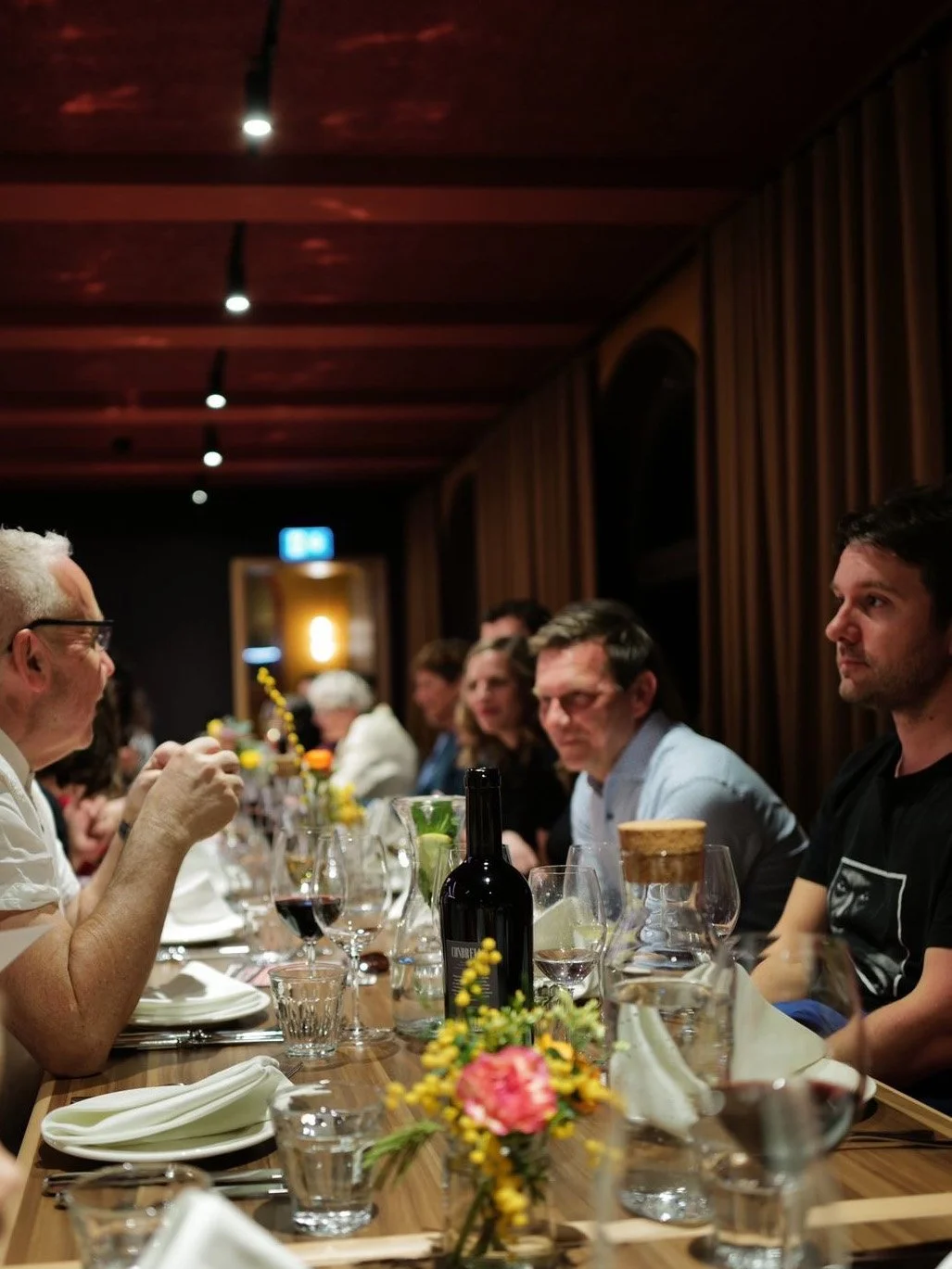 Menschen sitzen an einem festlich gedeckten Tisch in einem dunklen Raum mit Holzvertäfelung. Es gibt Blumen, Wein, Gläser und Teller auf dem Tisch, und die Atmosphäre wirkt gemütlich.