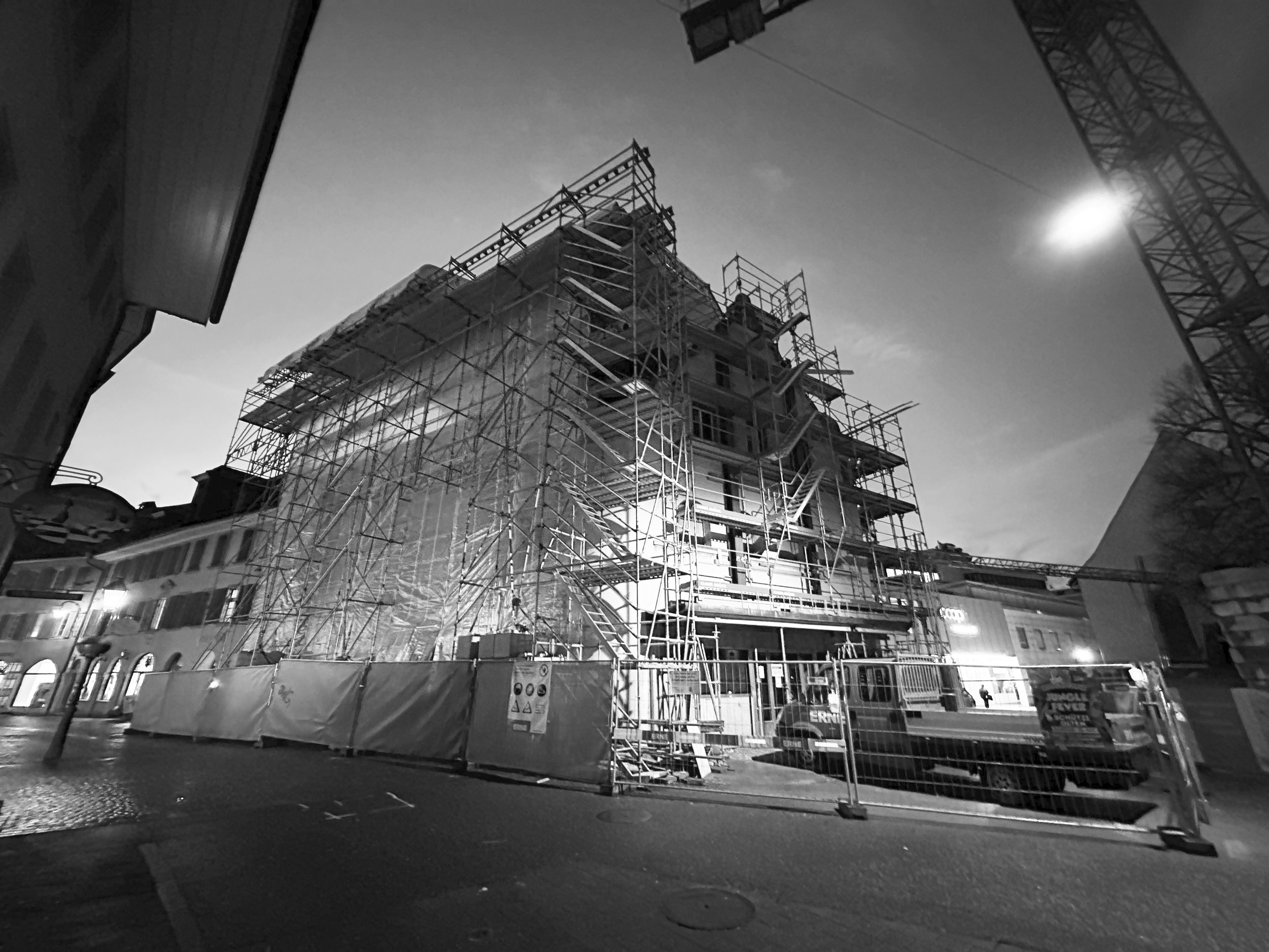 Mehrstöckiges Gebäude im Bau, umgeben von Gerüsten, in einer Stadt bei Nacht, mit beleuchteten Fenstern und Verkehrszeichen auf der Straße