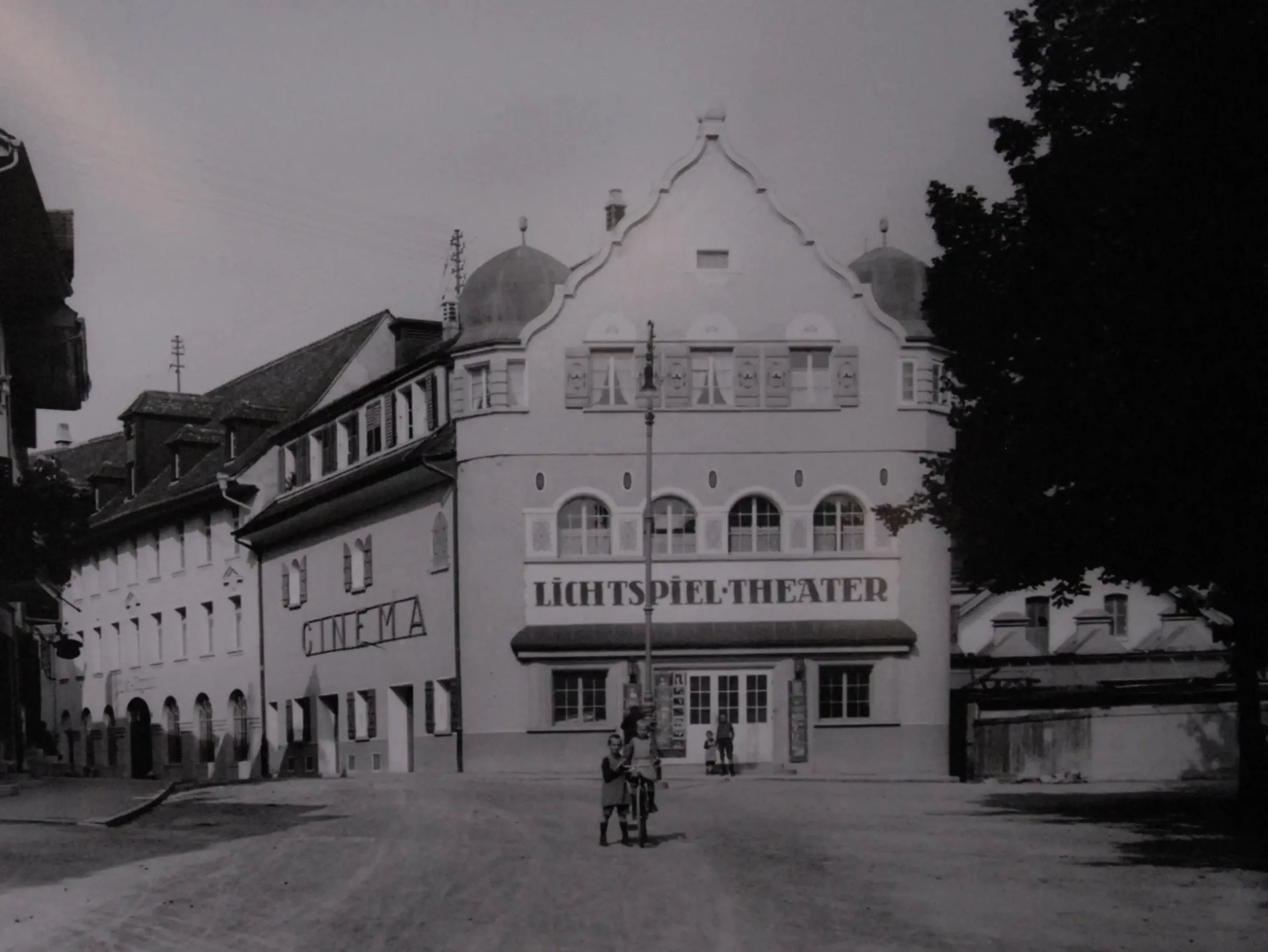 Ein altes Theatergebäude mit der Aufschrift "Lichtspieltheater" und "Kino" an der Fassade, Menschen stehen draußen und unterhalten sich.