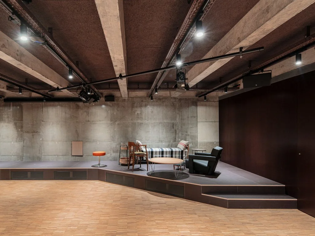 Ein leeres Theater oder Veranstaltungsraum mit Bühne, Stühlen und Sessel auf einer kleinen Plattform, unauffällig beleuchtet mit Scheinwerfern, mit Holzboden.