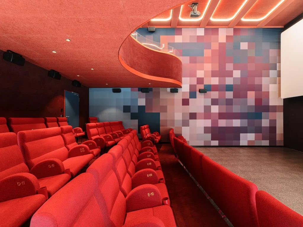 Leerer Kinosaal mit roten Sitzreihen, einer großen Leinwand und einer Wand mit einem pixeligen, bunten Muster.