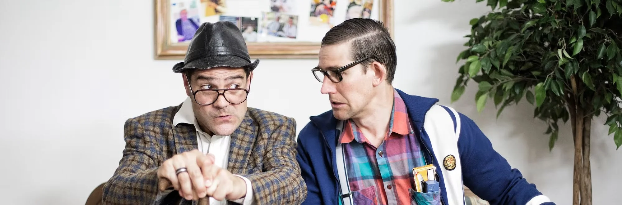 Zwei Männer sprechen sich an, einer trägt eine Brille, einen Hut und einen karierten Anzug, der andere trägt eine Brille, ein sauberes weißes Hemd, eine bunte Flanelljacke und eine ärztliche Tasche. Der Hintergrund ist eine Wand mit einem Bilderrahmen und eine Pflanze.