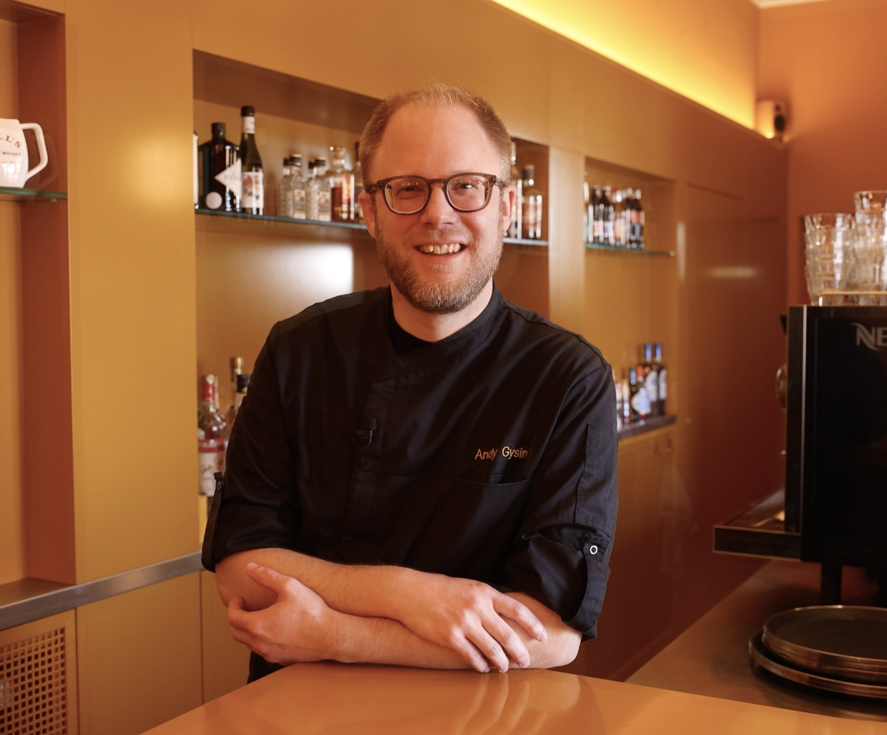 Ein männlicher Koch trägt eine schwarze Kochjacke mit dem Namen "Andy Gysin" und steht in einer modernen, warm beleuchteten Bar oder Restaurantküche, umgeben von Flaschen und Gläsern.