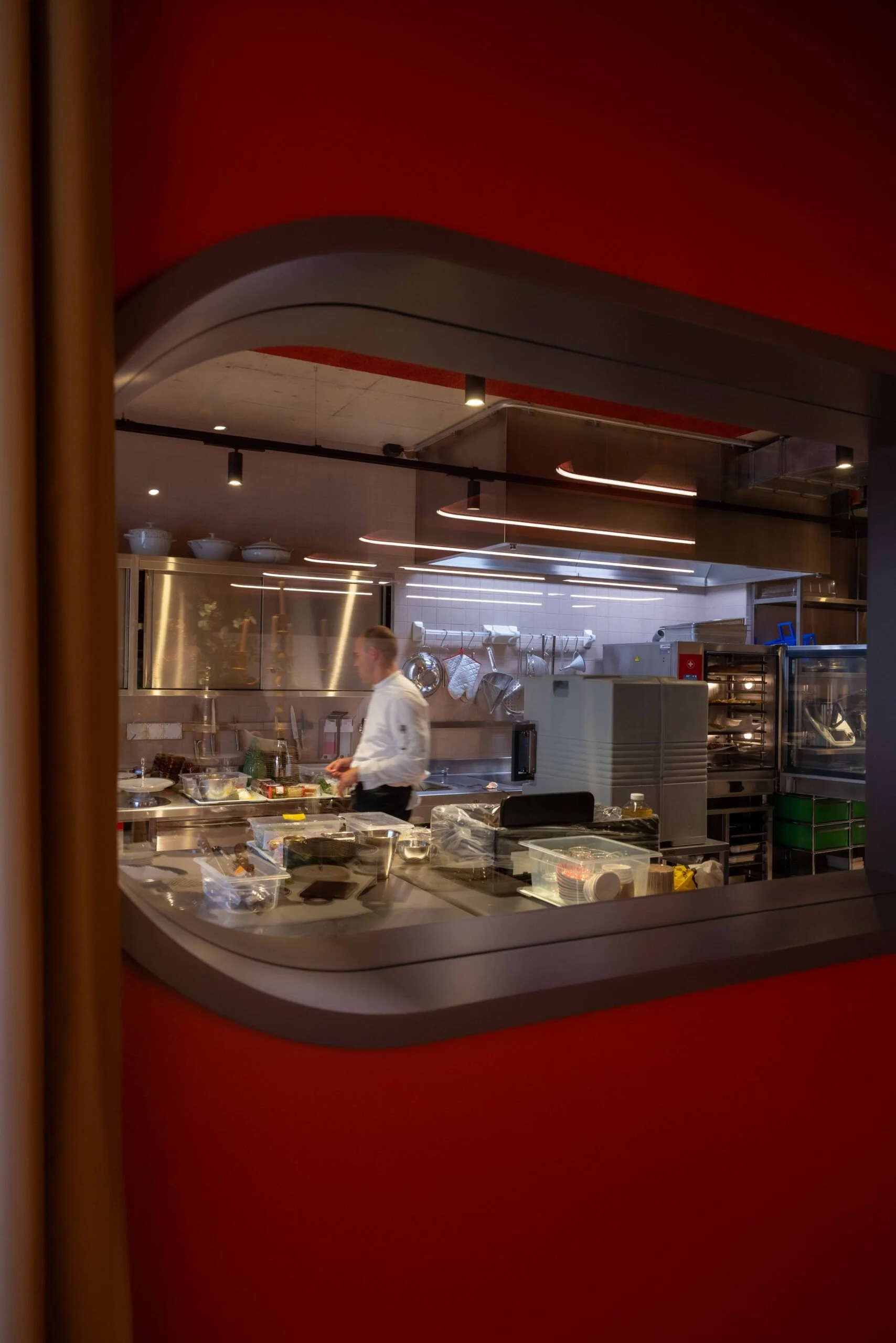Blick durch ein Fenster in eine Restaurantküche, mit einem Koch, der vor einem Arbeitsplatz mit verschiedenen Küchenutensilien steht.