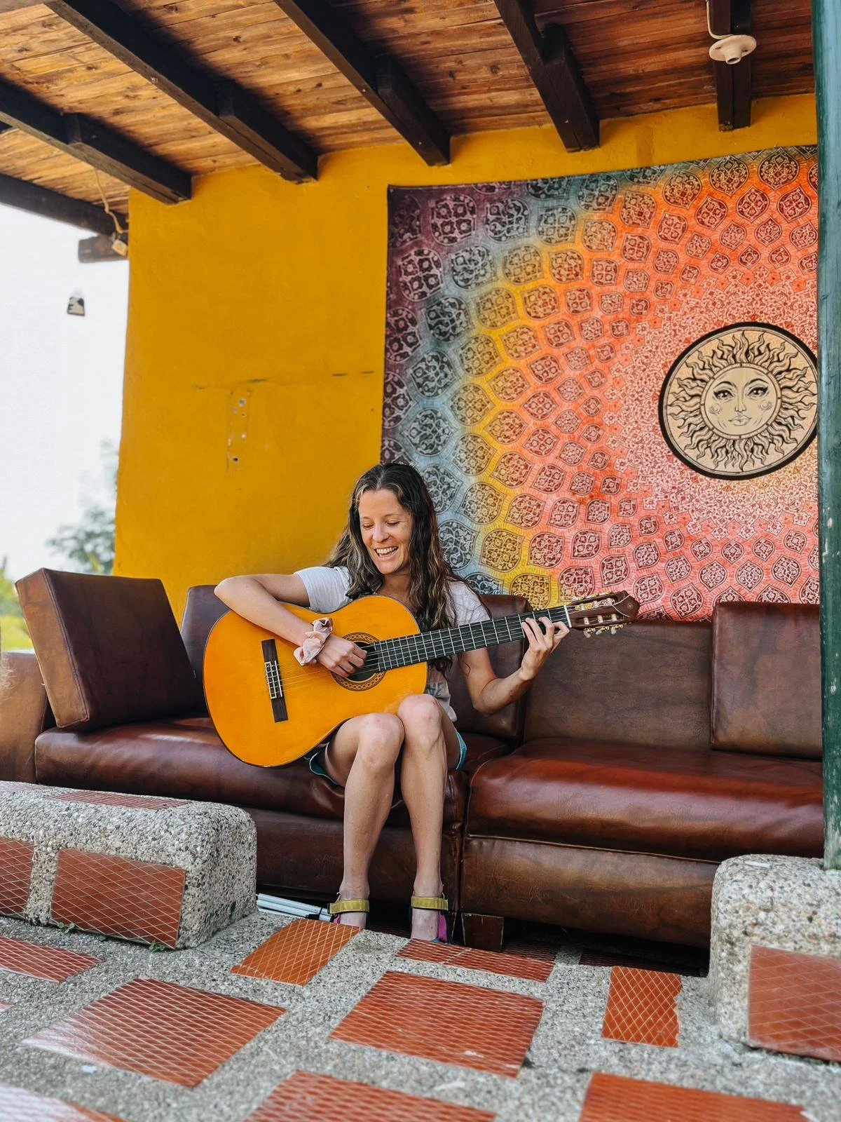 Ashley plays a guitar on an outdoor couch with a bright tapestry of colors and the sun behind her. She is radiating warmth, happiness, and a path forward.