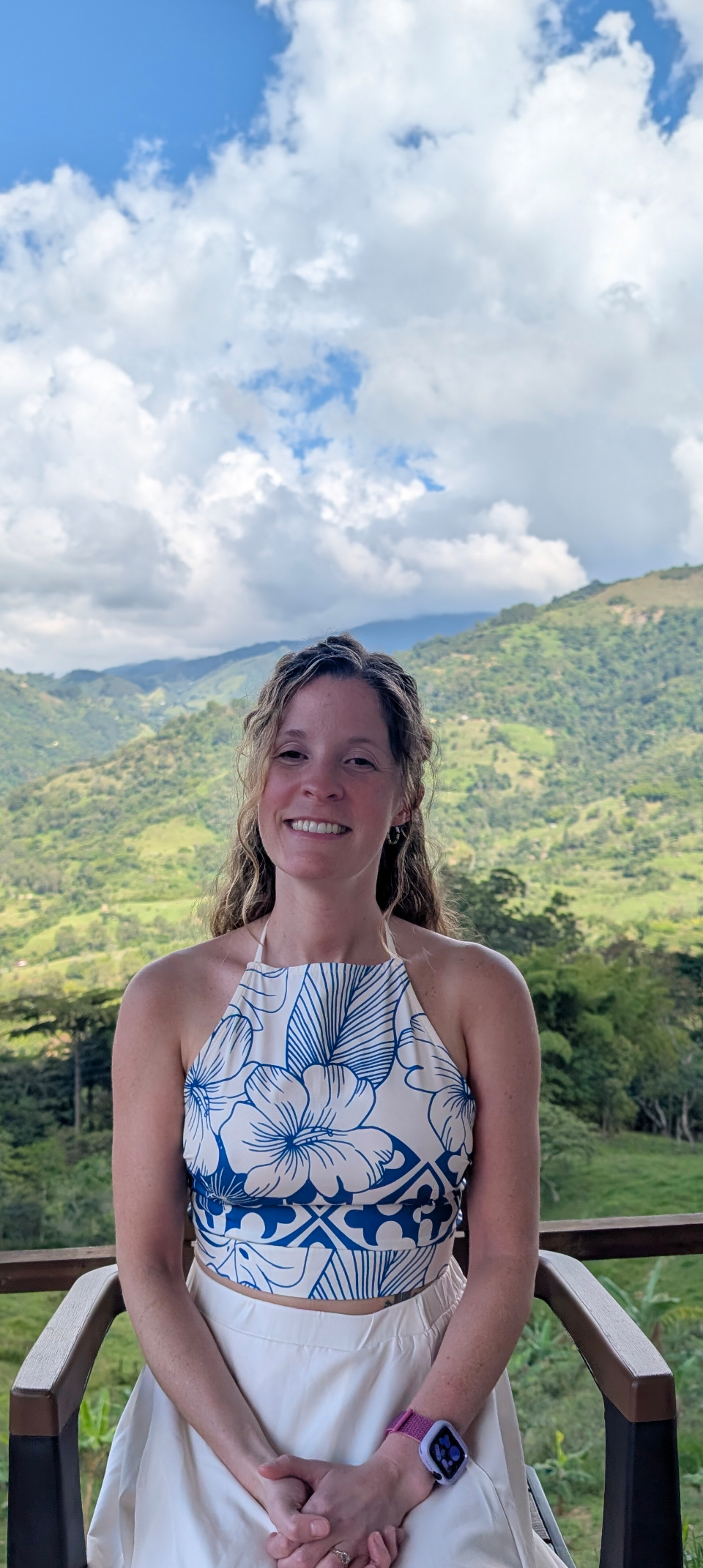 Denver transformational coach, Ashley Townsend, in a blue and white flower dress smiling and sitting with a beautiful green lush landscape behind her in the distance.