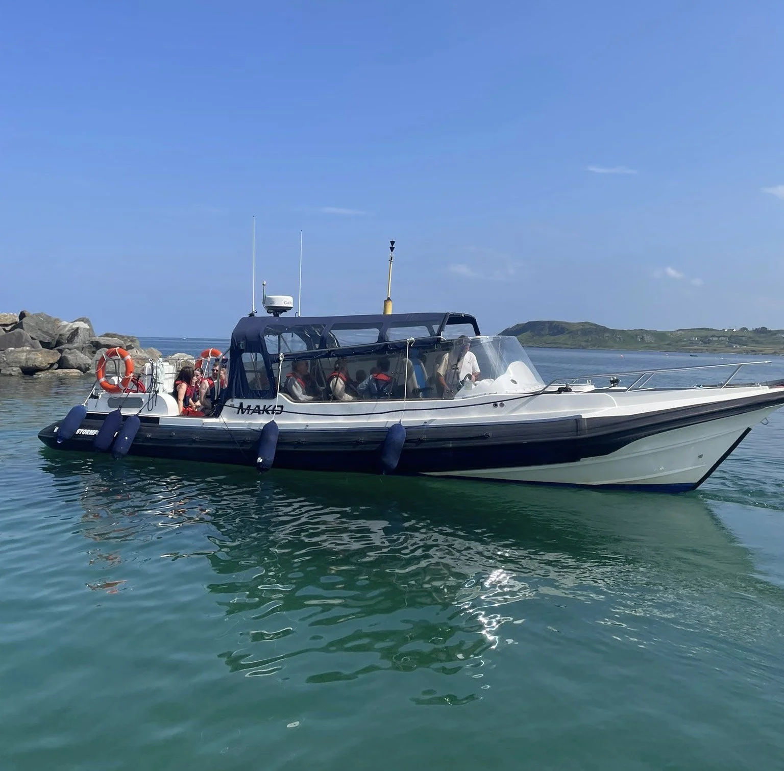 A black and white passenger boat named 'MAK' sailing in calm water near rocky shoreline under a blue sky with some clouds, with green hills in the background.