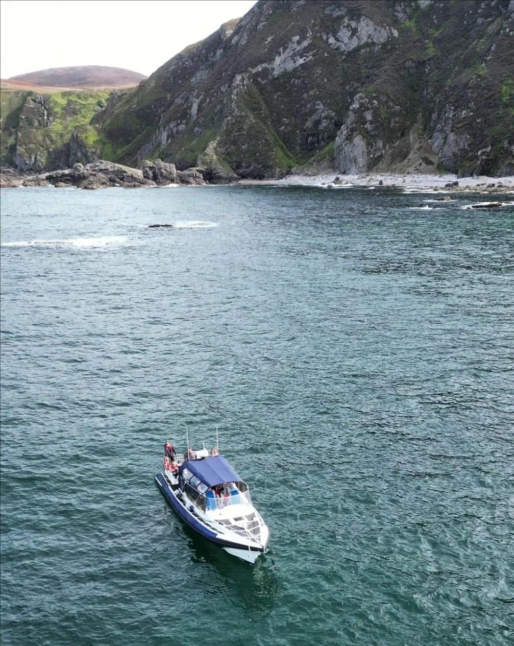 A boat with a blue canopy navigates through a body of water near rocky cliffs and green hills.